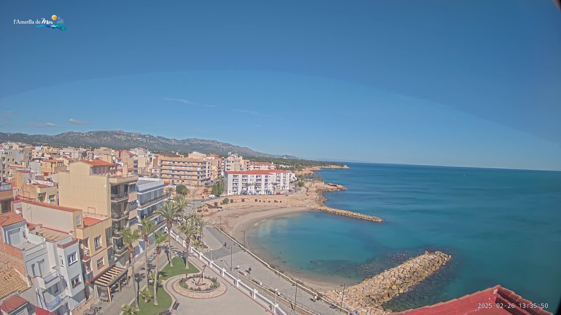 A coastal town with buildings and a small sandy beach is visible under a clear blue sky on a sunny day.
