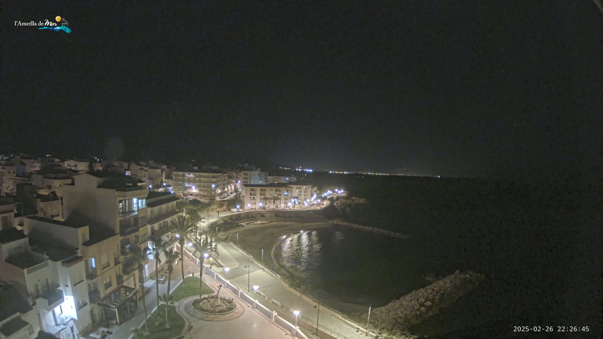 A nighttime view of a coastal town with buildings lit up along a curving shoreline and calm water under a clear, dark sky.