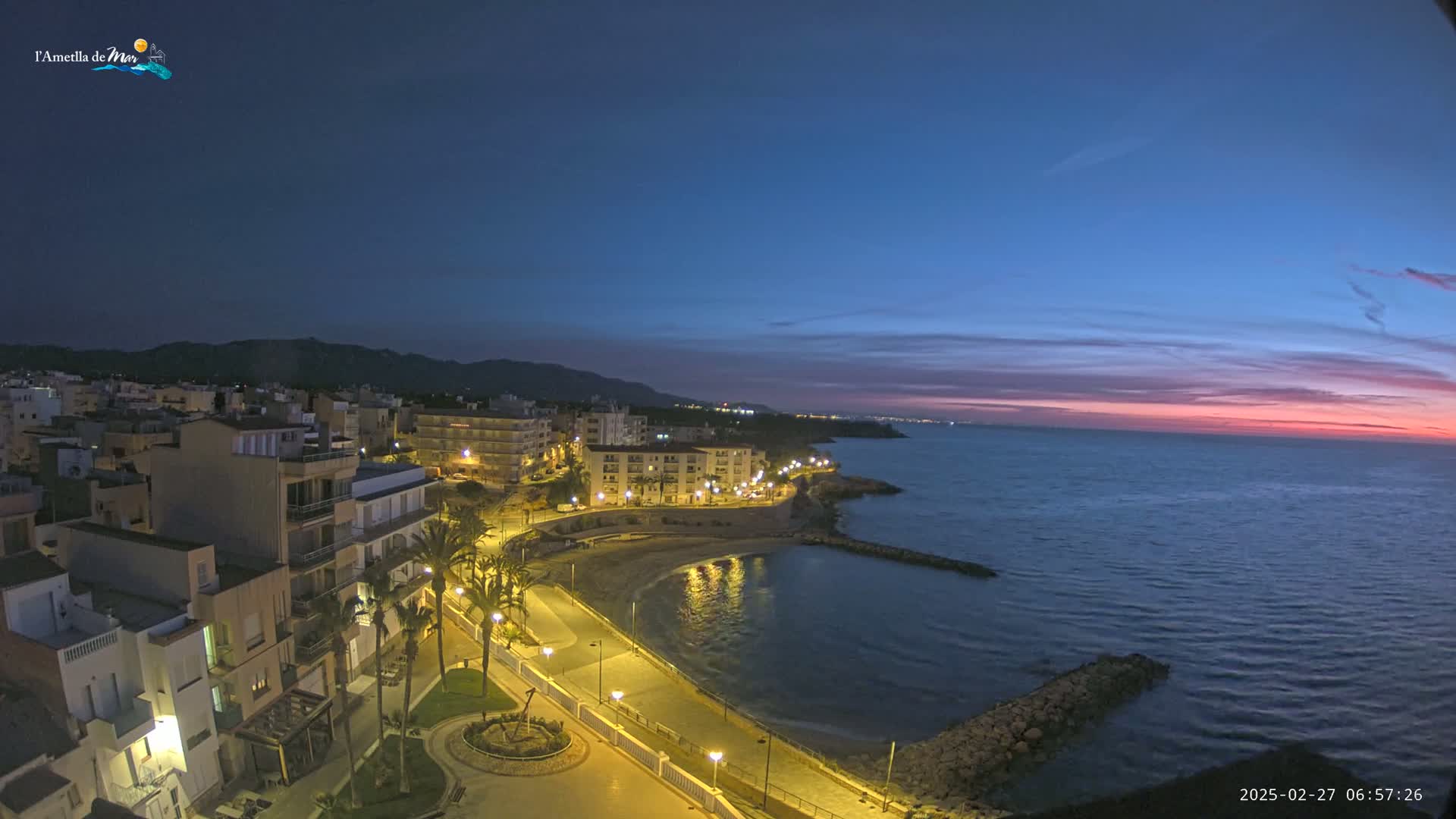 A coastal town is illuminated at twilight, with a calm sea reflecting the pink and orange hues of the sunset.
