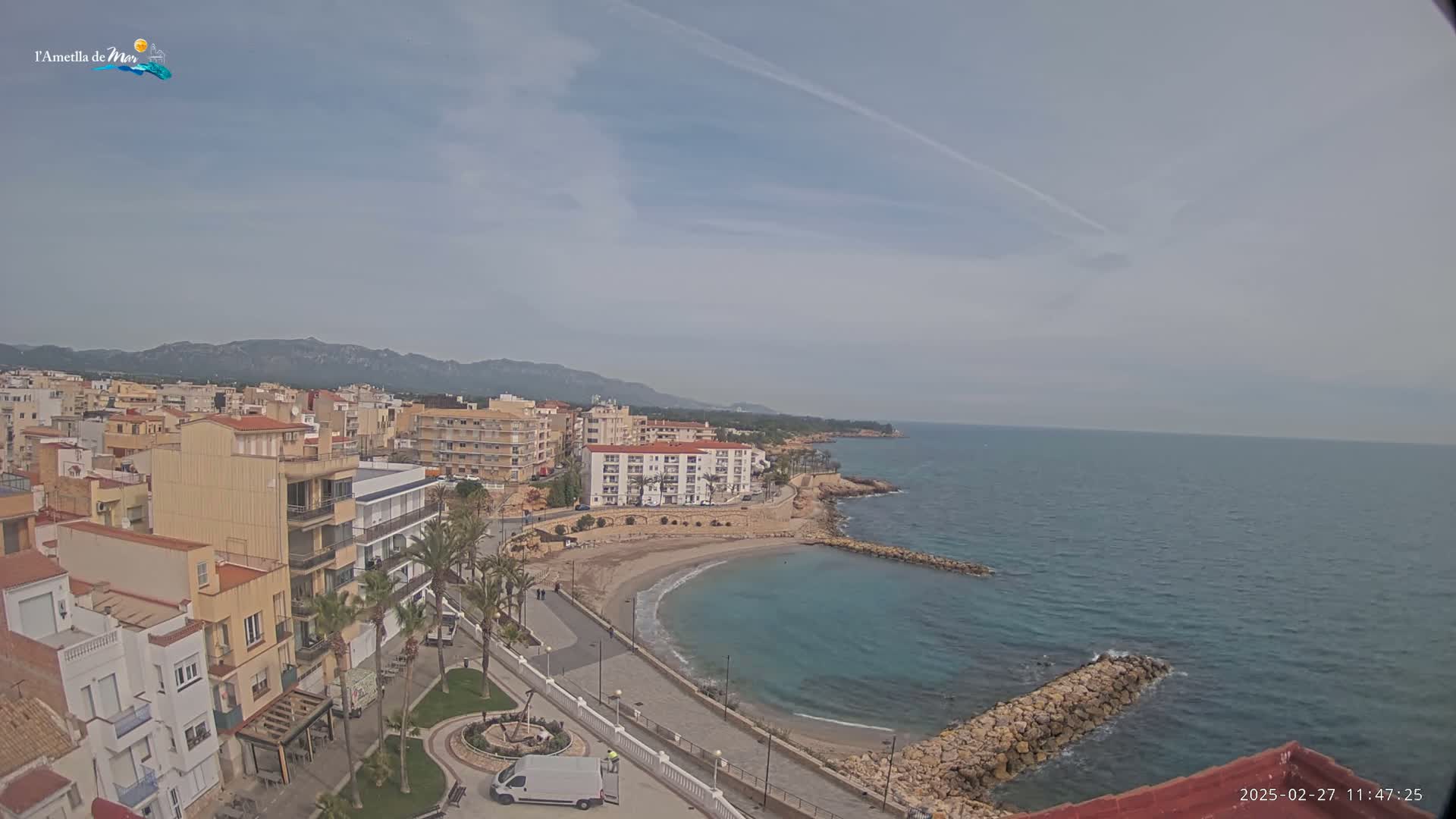 An aerial view shows a coastal town with buildings, a small sandy cove, and calm ocean under a mostly cloudy sky.