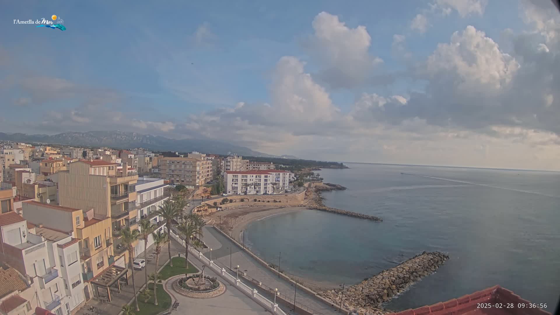 A partly cloudy day overlooks a coastal town with a small, sandy cove, buildings, and mountains in the distance.