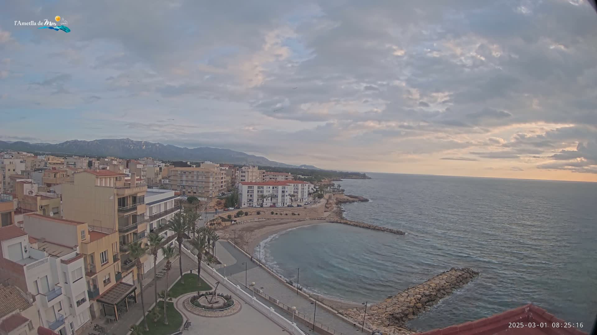 An elevated view shows a coastal town with buildings along a curving shoreline and calm sea under a cloudy sky.