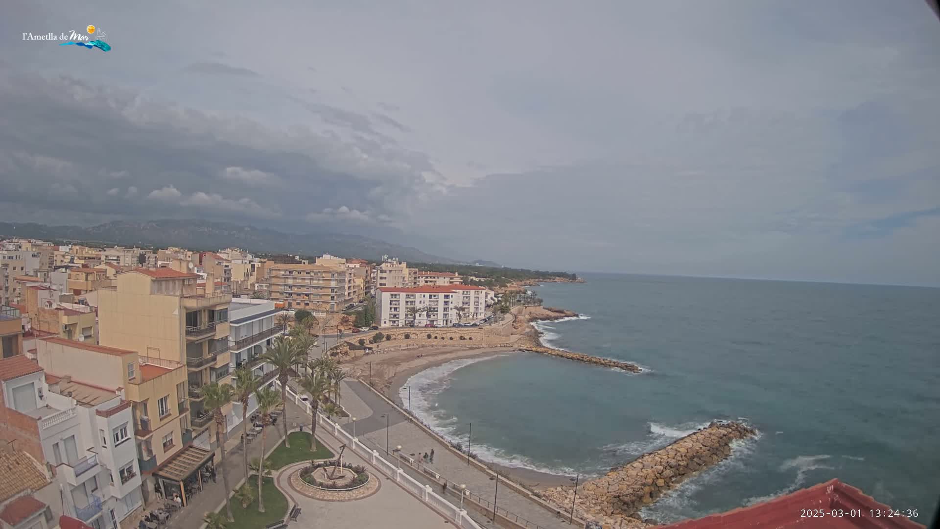 An elevated view of a coastal town on a partly cloudy day shows a small sandy beach curving into a calm sea protected by rock jetties, with buildings and palm trees lining the shore.
