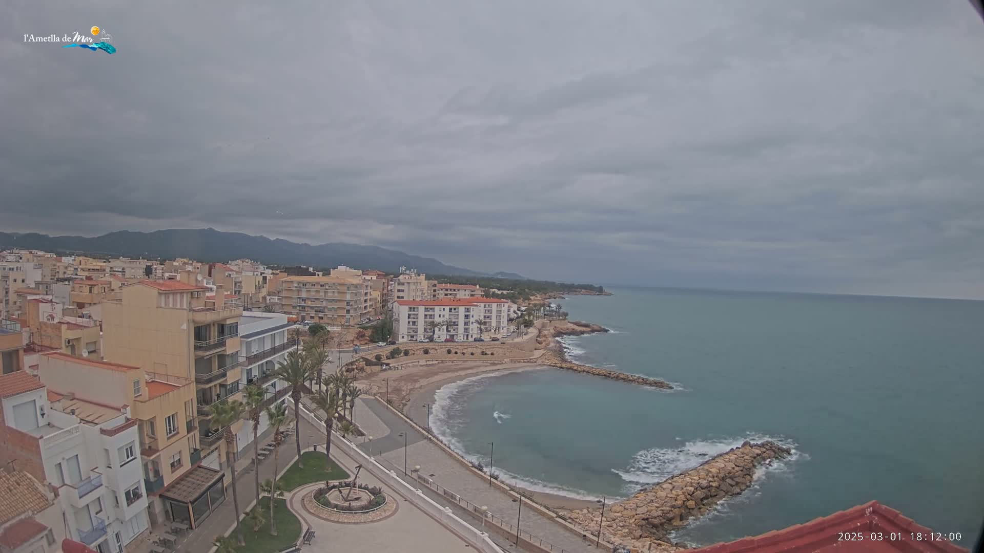 An overcast day reveals a coastal town with buildings, a small beach, and a calm sea partially enclosed by a rock jetty.