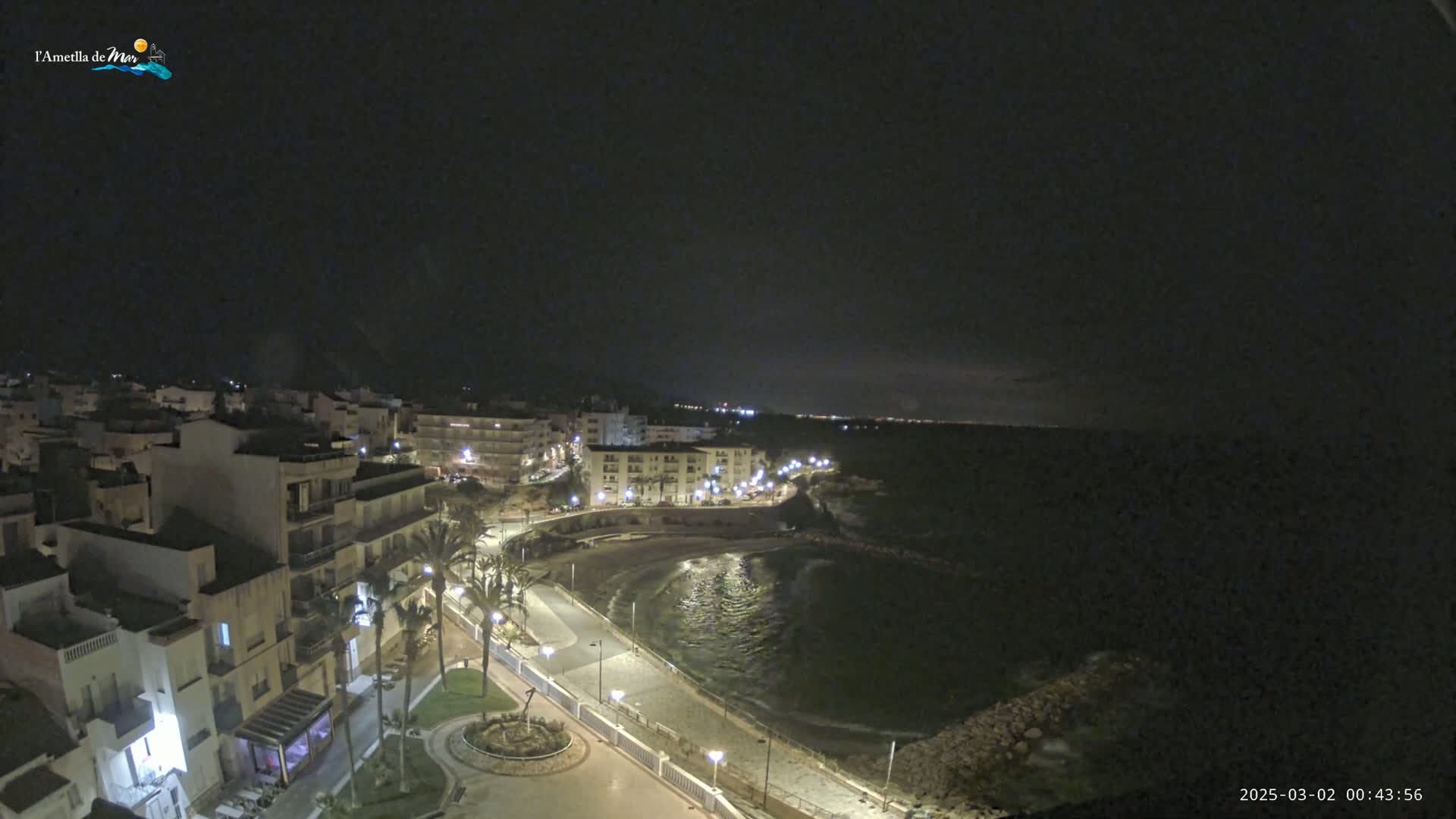 A nighttime coastal town with a calm bay, illuminated buildings, and palm trees along a paved waterfront path under a dark sky.