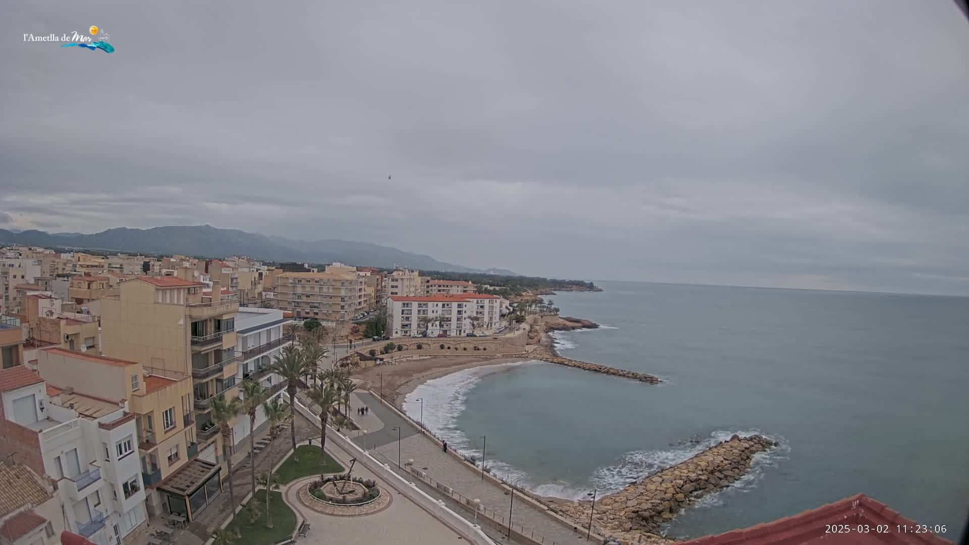A coastal town with buildings and a small beach is visible under an overcast sky, with calm waves lapping at a rocky shore and mountains in the background.