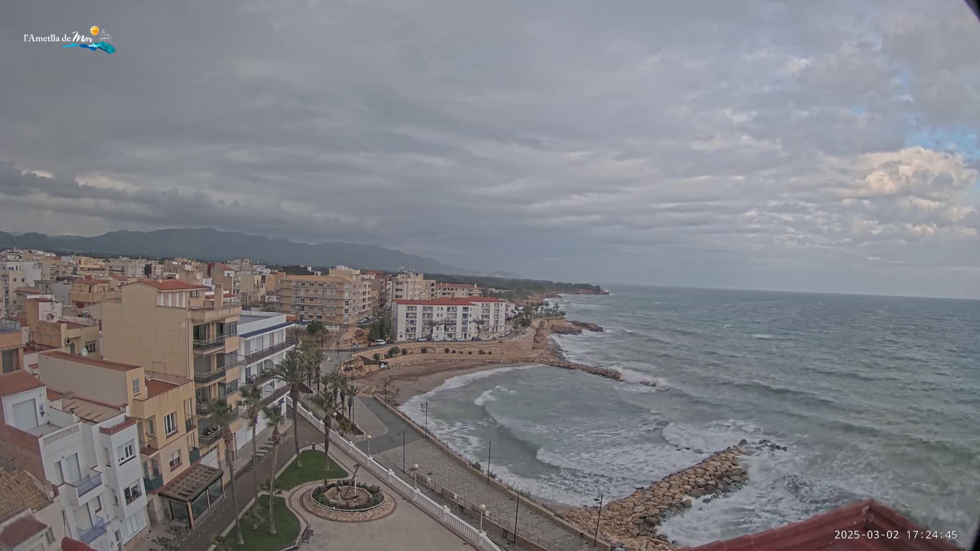 A coastal town with low buildings and palm trees sits under an overcast sky, overlooking a wavy ocean with a small, calm cove.