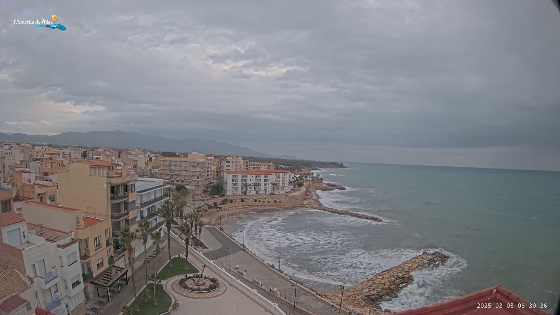 A coastal town with buildings and palm trees overlooks a rocky shore and a wavy sea under an overcast sky.