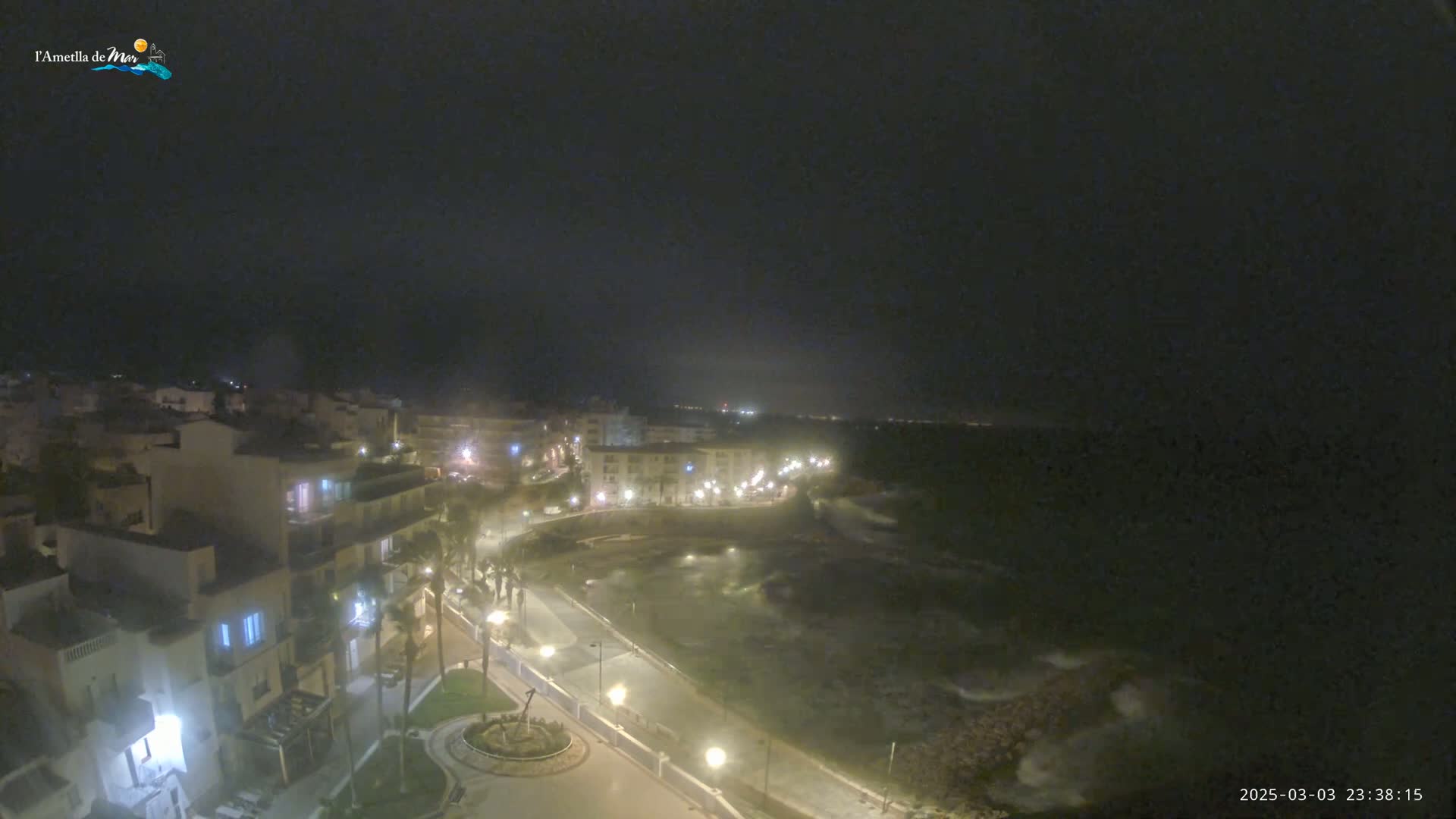 A nighttime view of a coastal town with a paved walkway overlooking a dark, wavy ocean under a cloudy sky.
