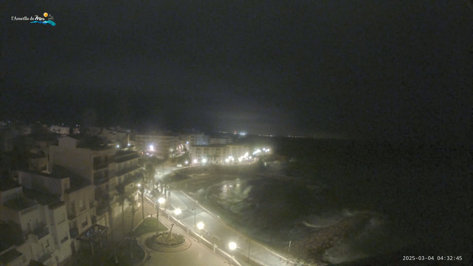 A nighttime, high-angle view shows a coastal town with buildings lit up along a paved walkway that curves around a dark body of water with small waves under a mostly clear, dark sky.