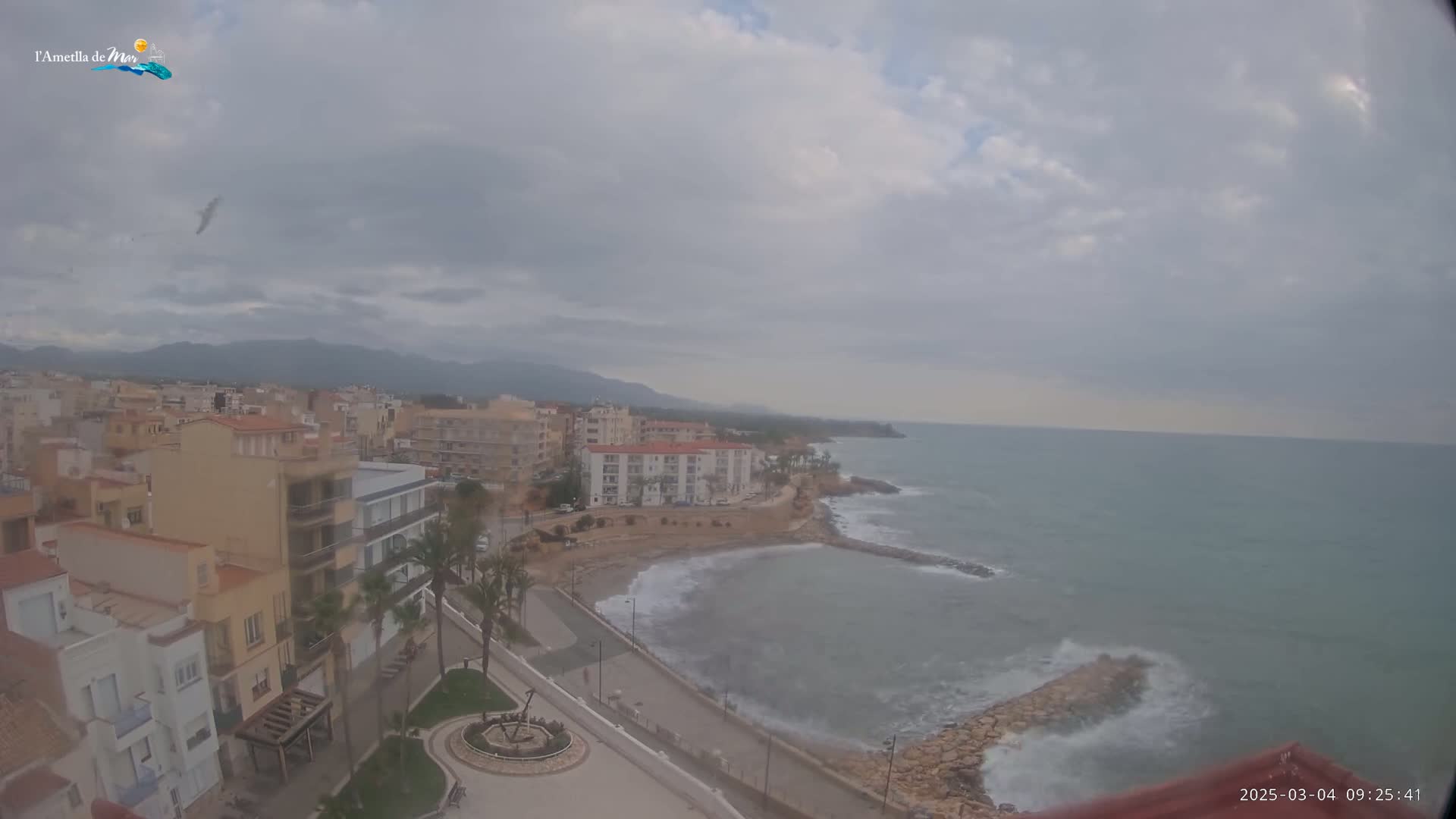 An overcast sky hangs over a coastal town with buildings, a shoreline, and a partly-calm sea with some small waves.