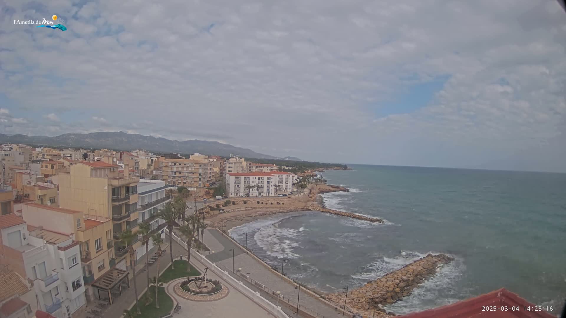 A coastal town with buildings and palm trees stretches along a rocky shoreline where small waves meet a calm sea under a mostly cloudy sky.