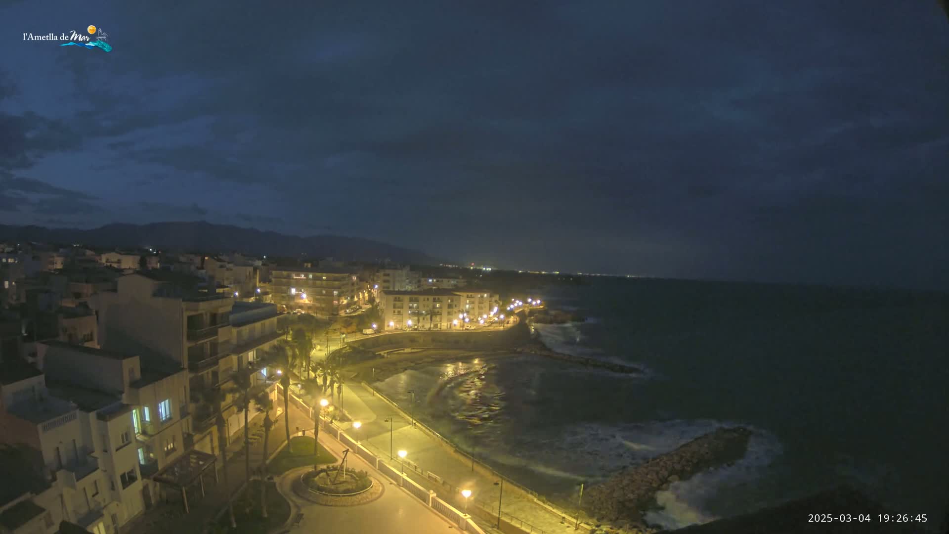 A nighttime coastal town with dimly lit buildings, palm trees, and a paved walkway along the dark ocean under a mostly clear, dark sky.