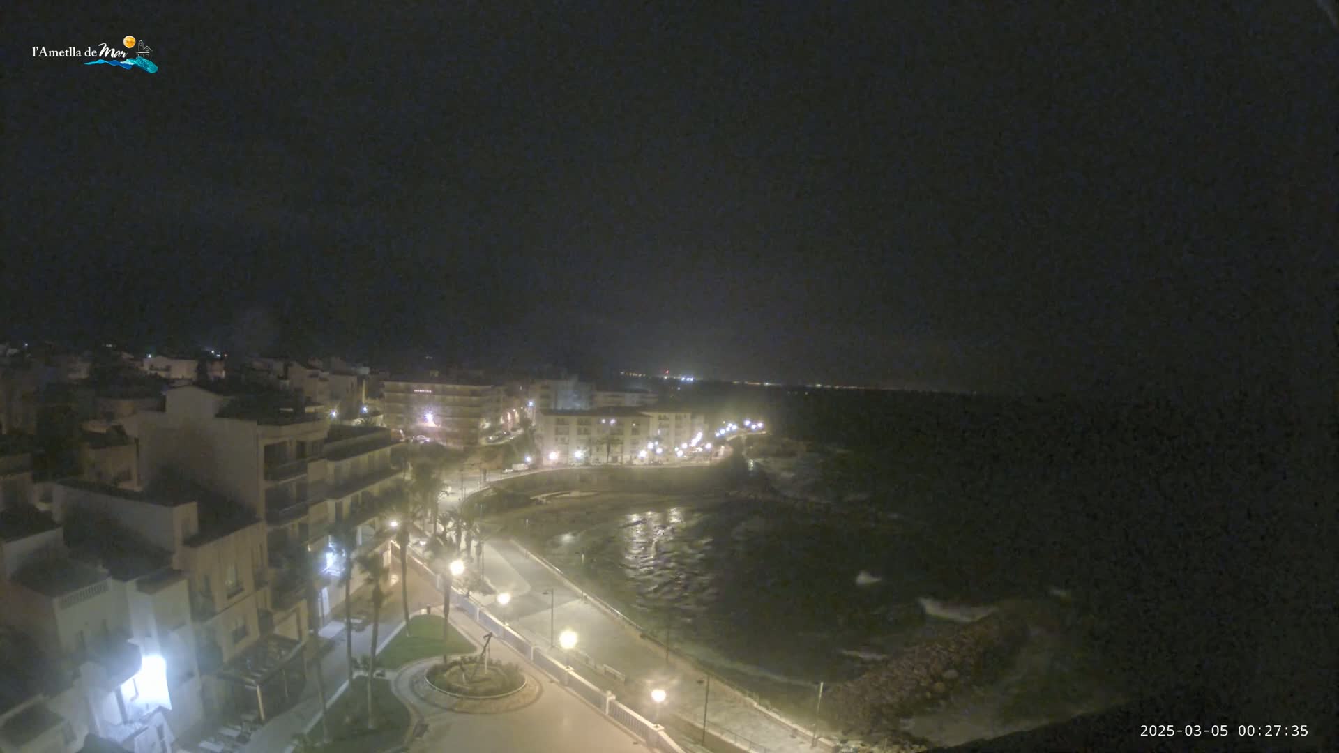 A nighttime, high-angle view shows a coastal town with buildings lit up along a curving road next to a dark, wavy ocean under a clear, dark sky.