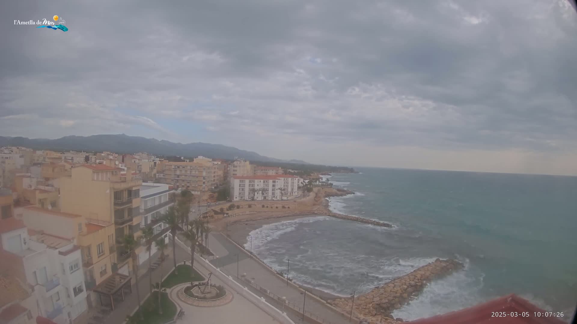 An overcast day reveals a coastal town with buildings, palm trees, a small beach, a rocky shoreline, and a choppy sea under a cloudy sky.