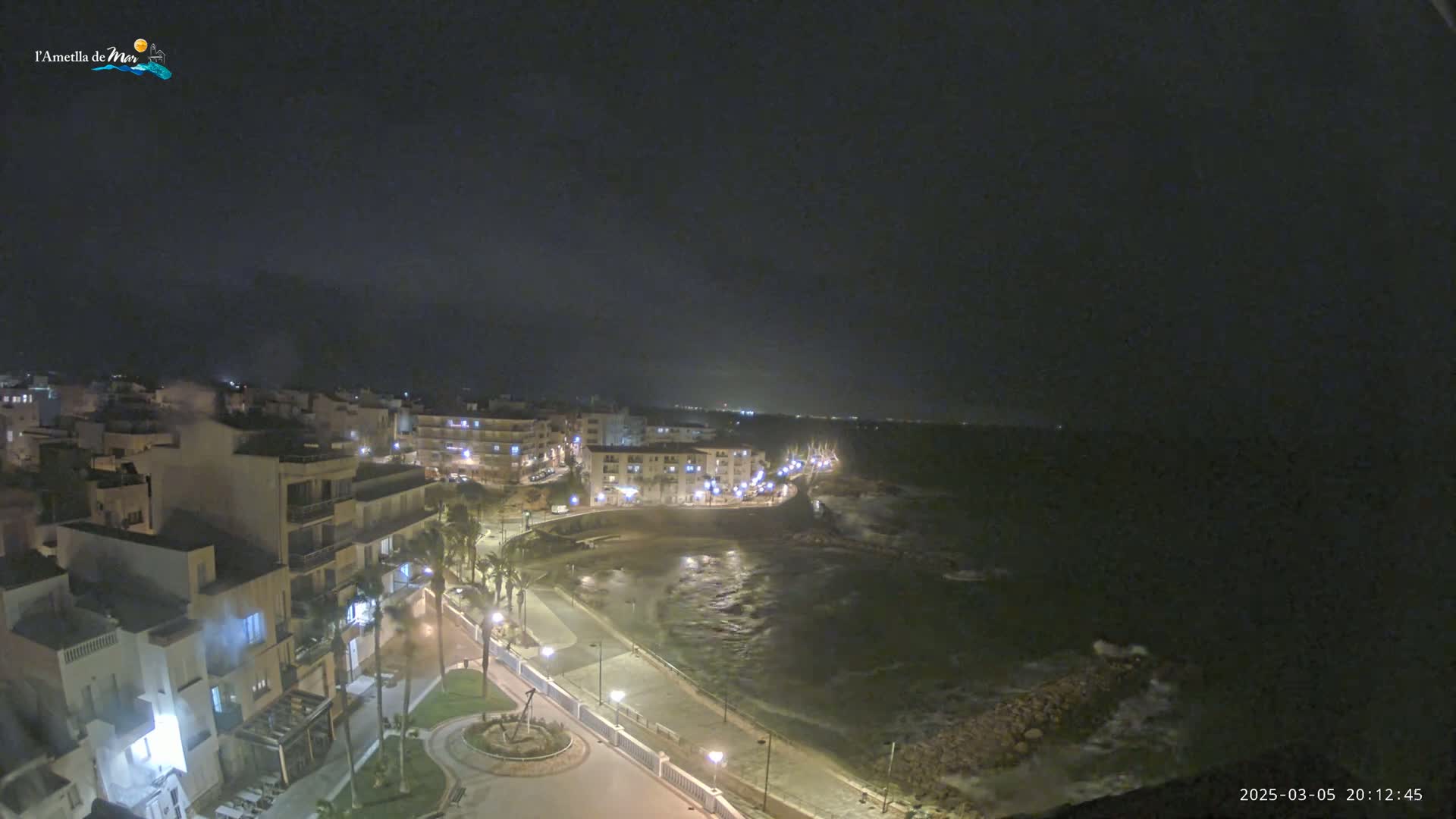 A nighttime, coastal town with illuminated buildings and a shoreline with dark, wavy water under a mostly clear, dark sky.
