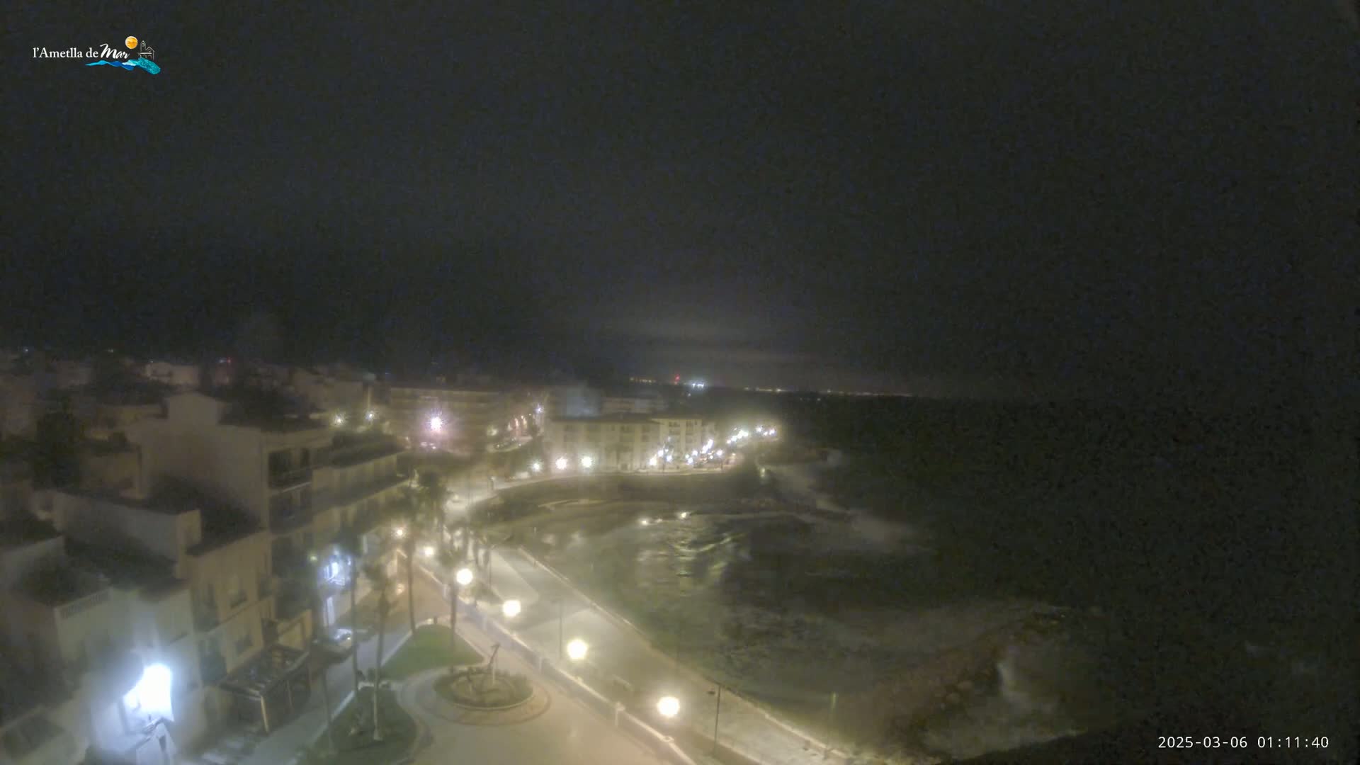A nighttime, high-angle view shows a coastal town with buildings illuminated by streetlights, a dark body of water, and a mostly cloudy sky.