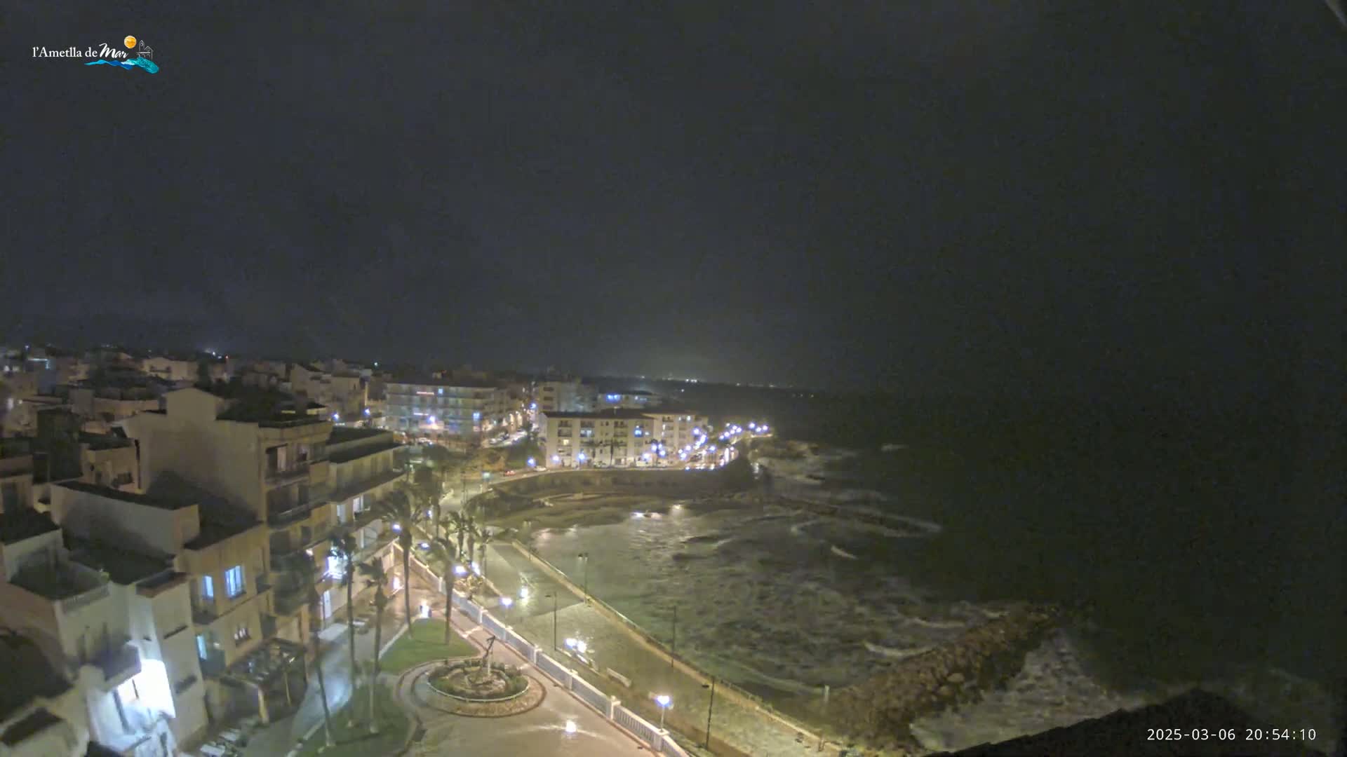 A nighttime coastal town is shown with moderately rough waves crashing against the shore under a dark sky.