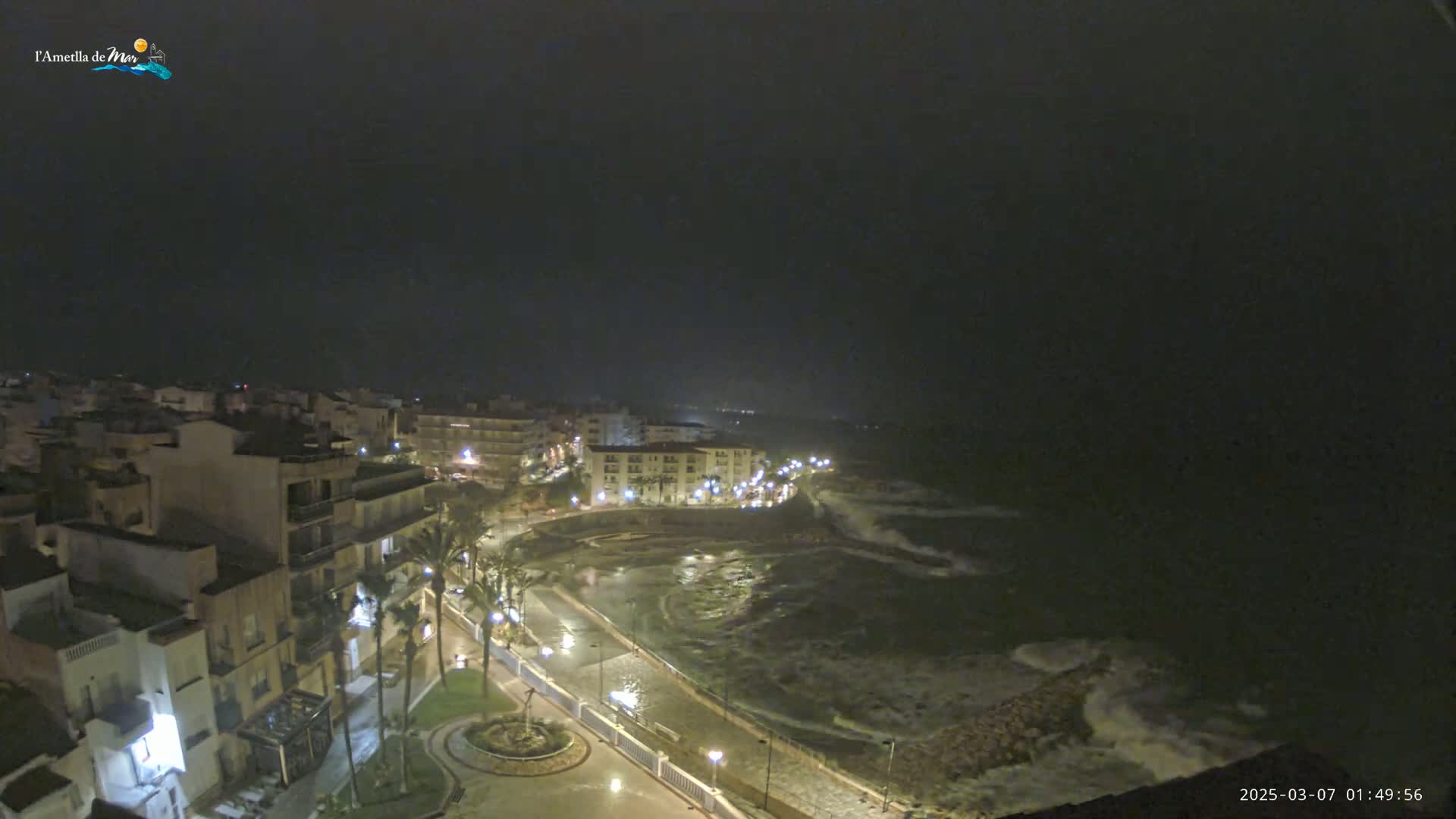 A nighttime coastal town is shown with moderately rough waves crashing against a seawall under a dark, overcast sky.