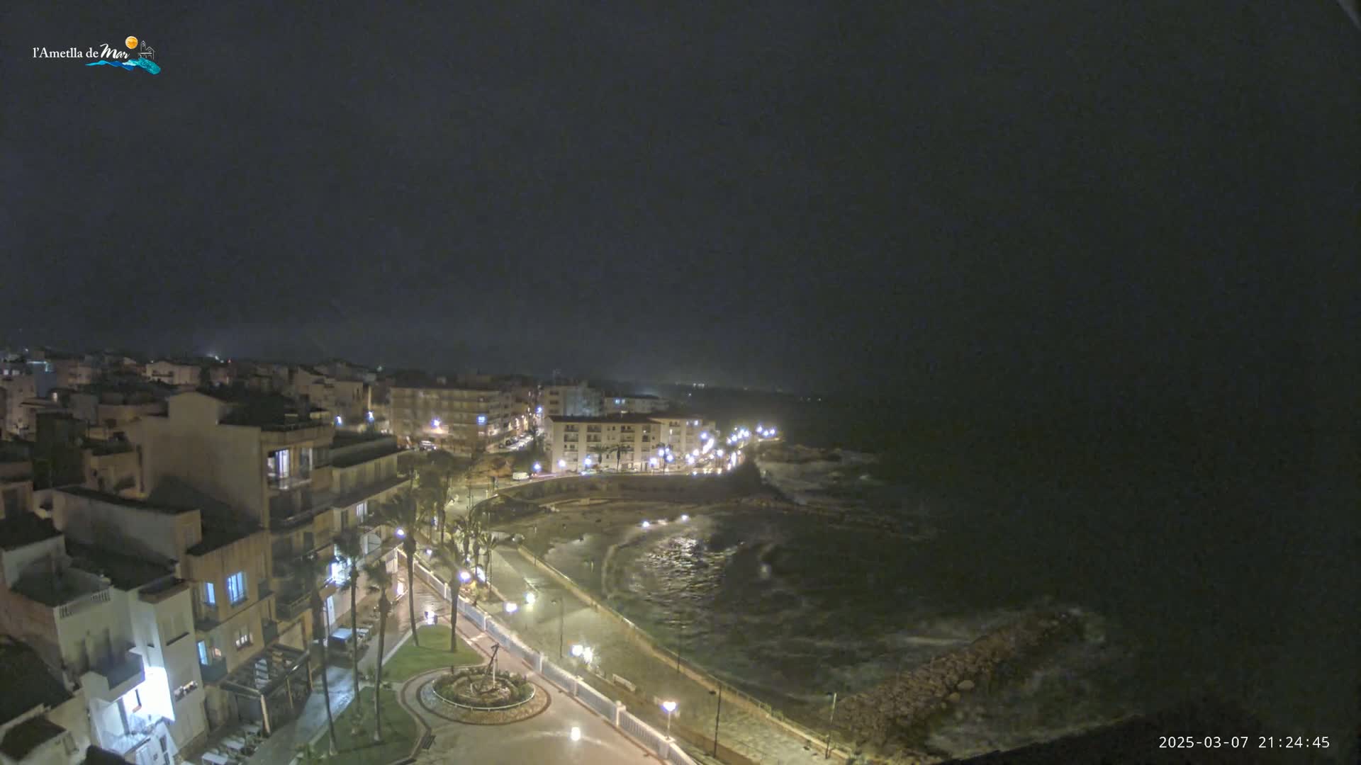 A nighttime, overcast view of a coastal town with a dark, wavy ocean and illuminated streets and buildings.