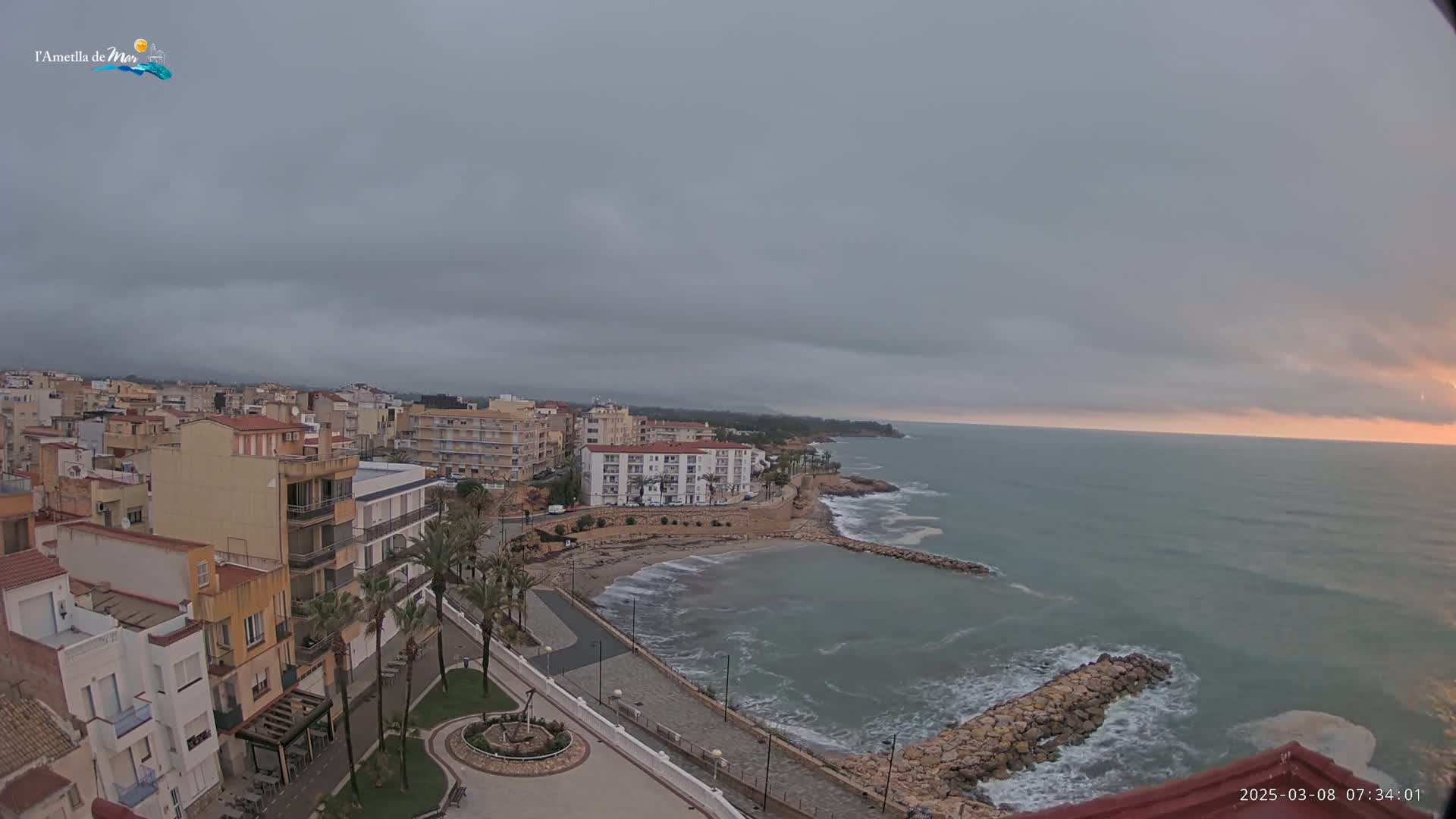 A coastal town under an overcast sky, with a calm sea meeting a small, sandy beach and a rocky breakwater.