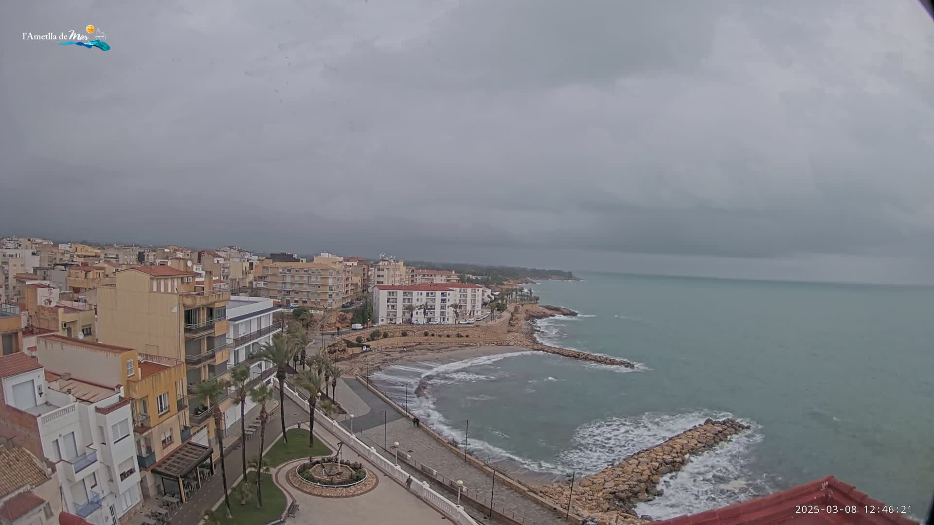 An overcast day shows a coastal town with buildings, palm trees, a small beach, and a rocky shoreline extending into a calm sea.
