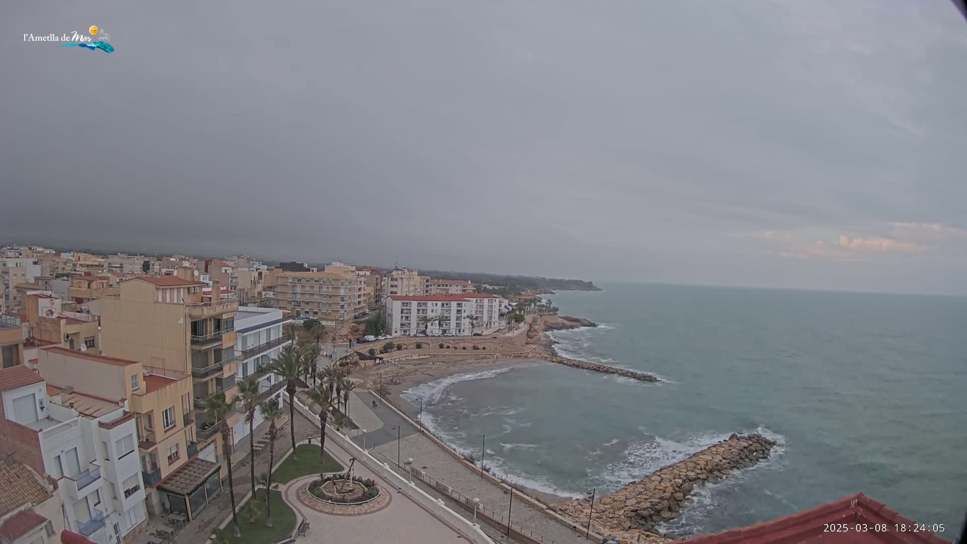 A coastal town under an overcast sky is seen from an elevated vantage point, showing a small beach and calm sea with some waves breaking against a rock jetty.