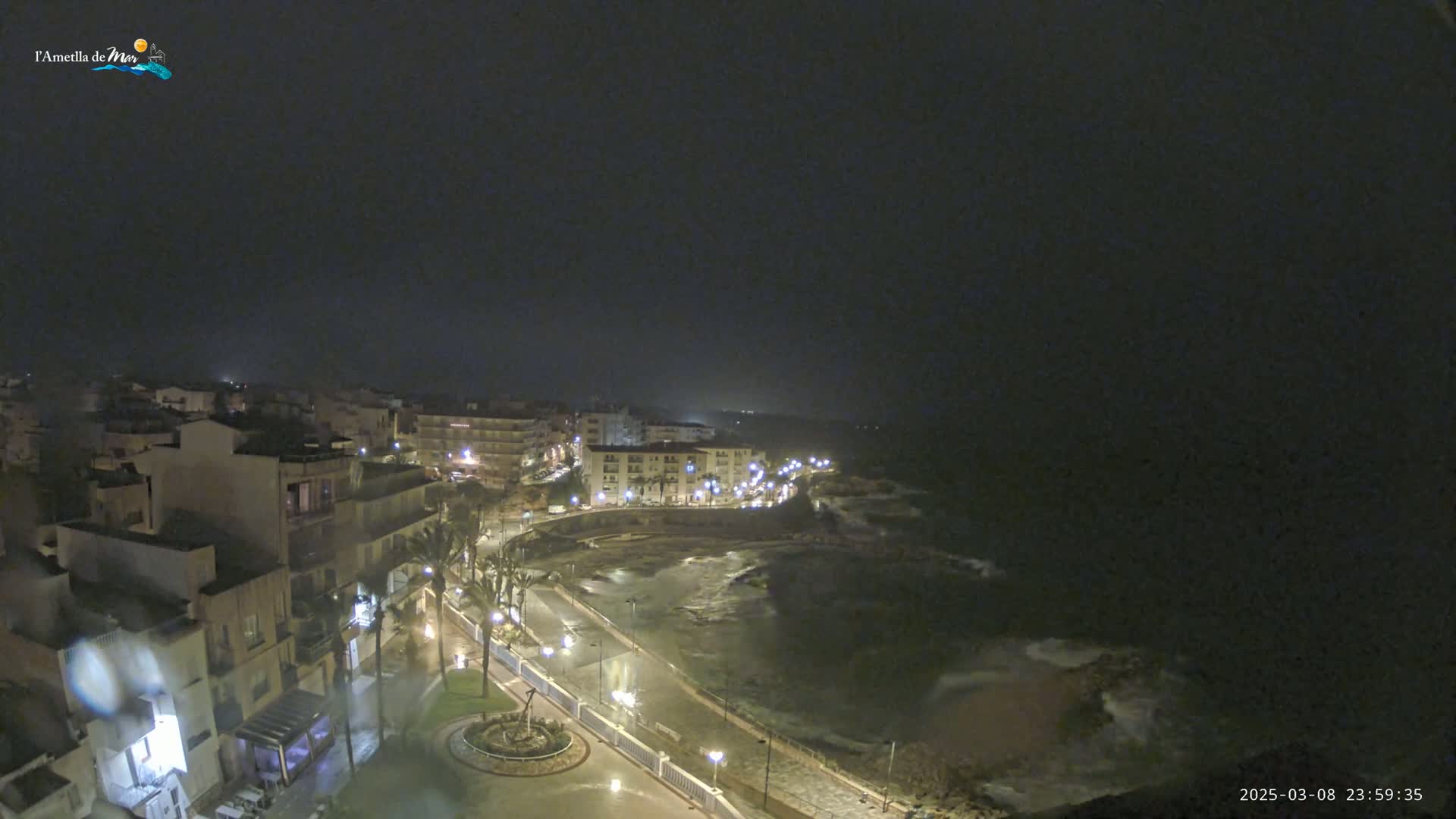 A nighttime coastal town is seen with rain, showing illuminated buildings, streets, and a dark ocean with some wave action.