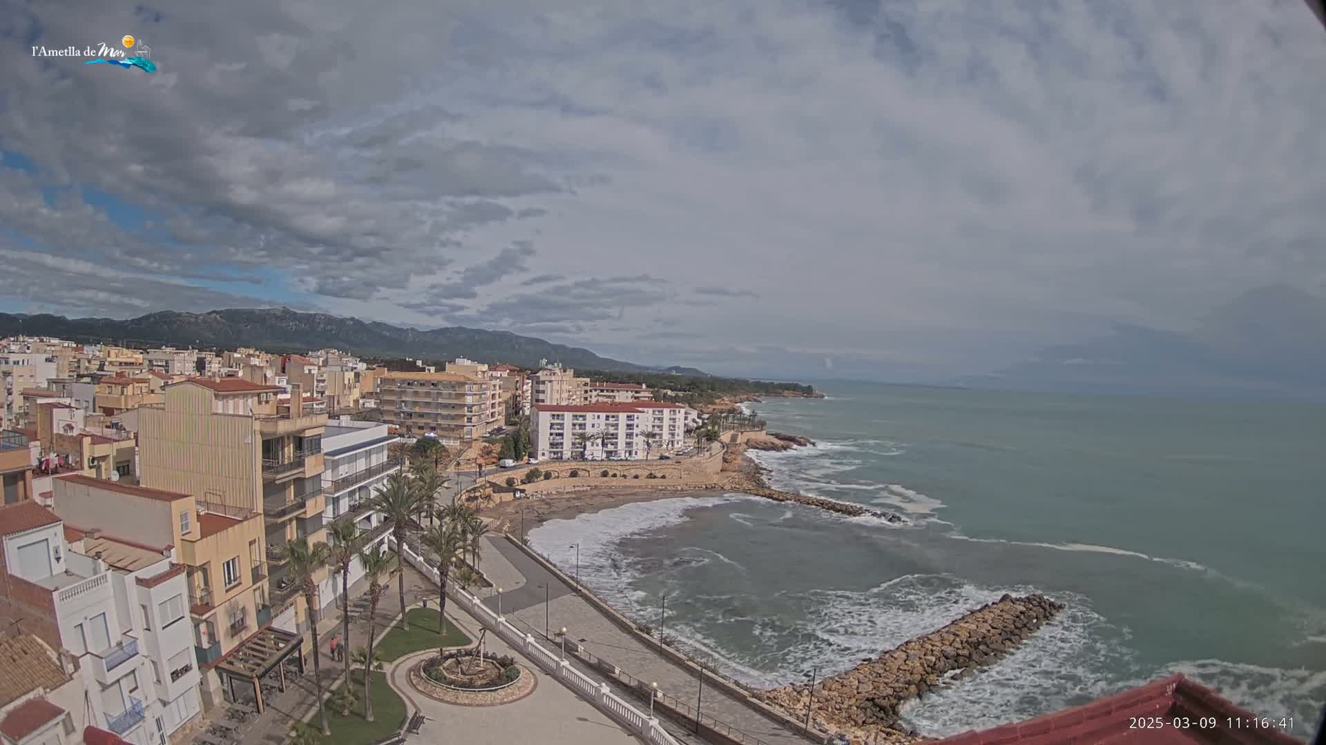A coastal town with buildings and palm trees overlooks a beach with moderate waves under a cloudy sky.