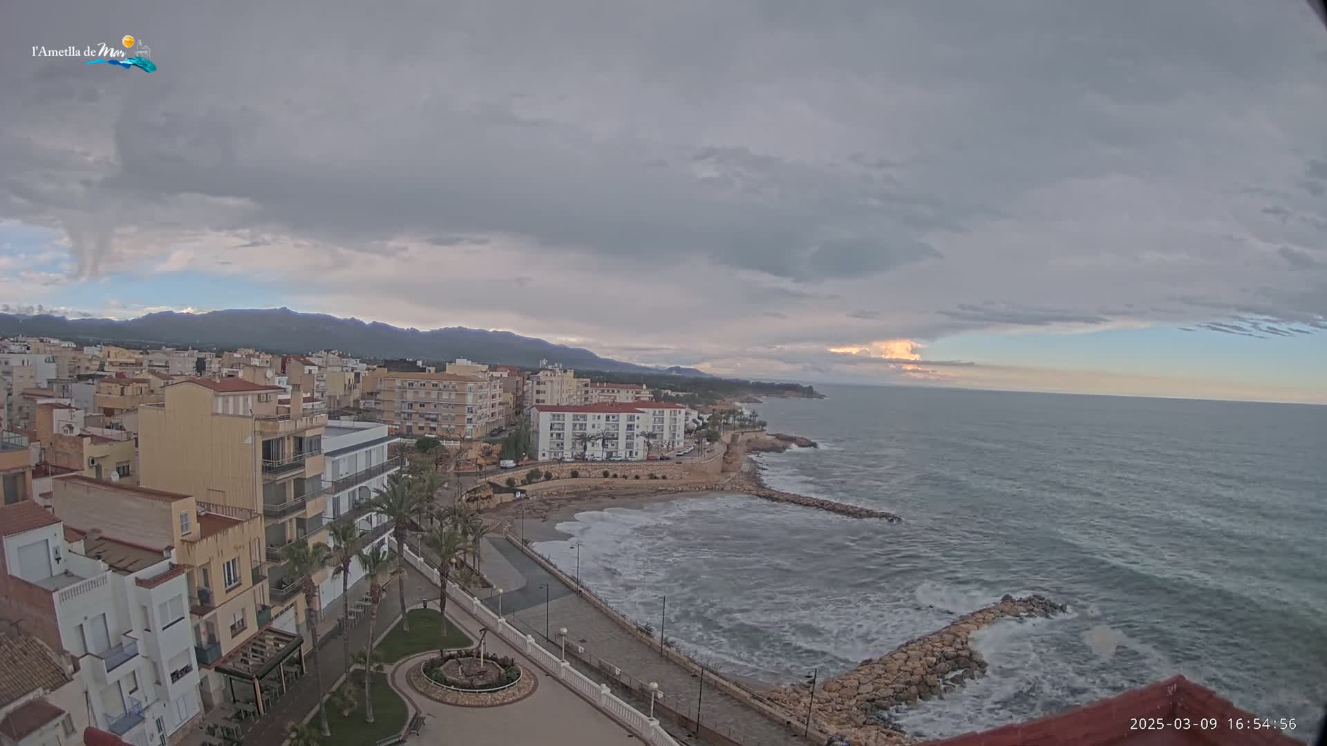 A coastal town with buildings and palm trees sits under a mostly cloudy sky, overlooking a sea with moderate waves crashing against a rocky shore.