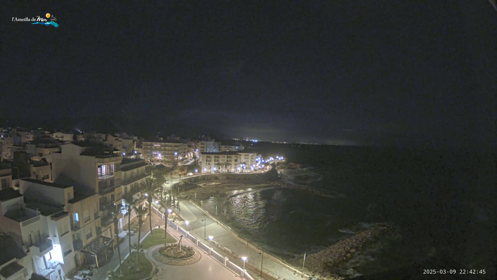 A nighttime view of a coastal town with lit buildings and a calm, dark ocean under a clear sky.