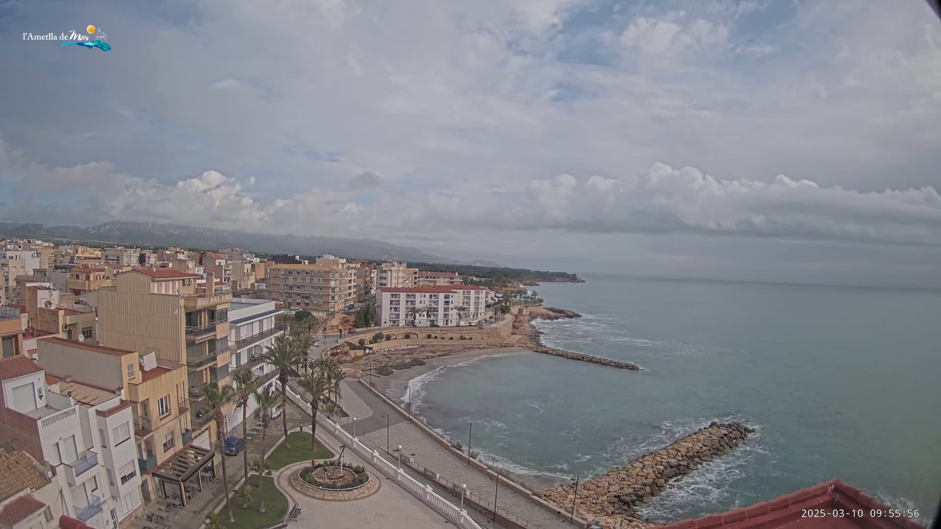 A coastal town with multi-story buildings sits on a partly cloudy day next to a calm ocean with small waves breaking against a rock jetty.