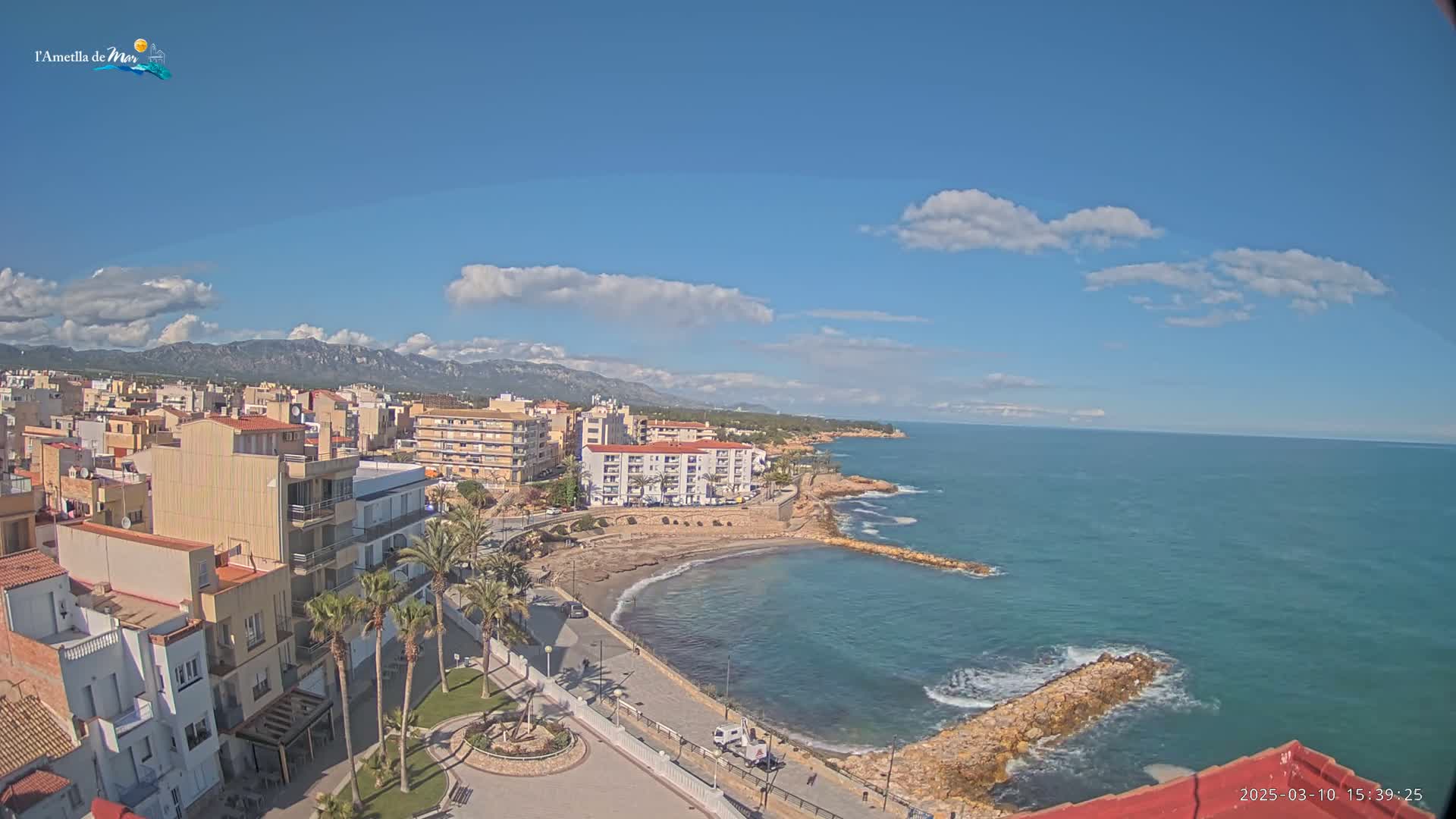 An elevated view shows a coastal town on a partly sunny day with a calm sea and mountains in the background.