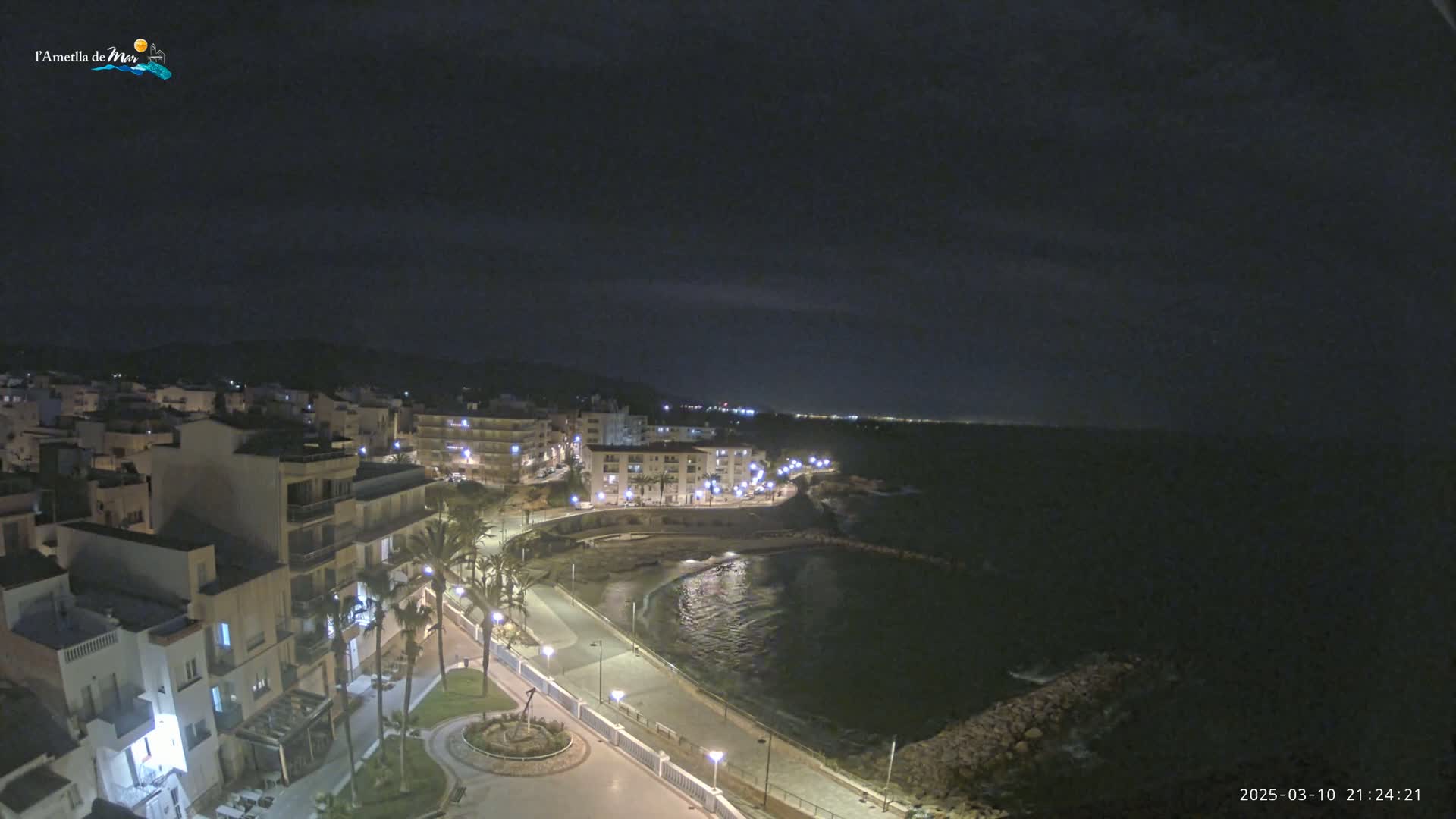 A nighttime aerial view of a coastal town with calm water, illuminated buildings, and palm trees lining a waterfront promenade under a dark, cloudy sky.