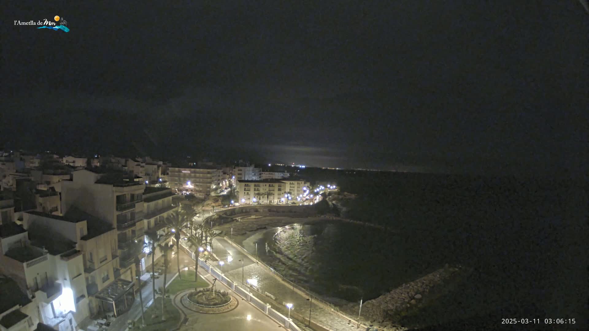 A nighttime, coastal town scene shows a dark body of water next to a paved walkway lined with palm trees and buildings with illuminated windows.