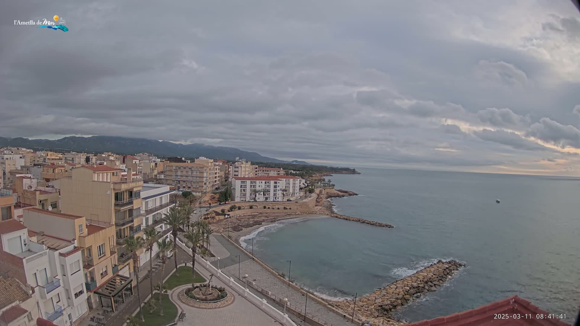 A coastal town with low buildings, palm trees, and a small sandy beach is nestled beneath a cloudy sky overlooking a calm sea and distant mountains.