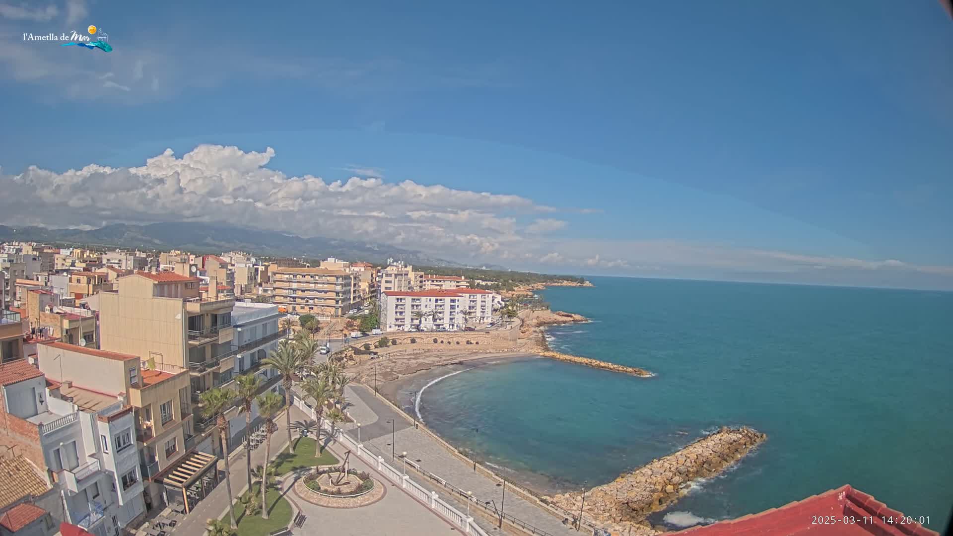 A coastal town with buildings, palm trees, and a small, curved beach is visible under a partly cloudy sky, with a calm sea extending to the horizon.