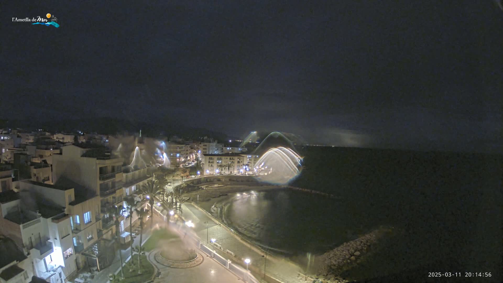 A nighttime coastal town is illuminated, with a calm body of water reflecting light and what appears to be a water feature creating arched light trails.