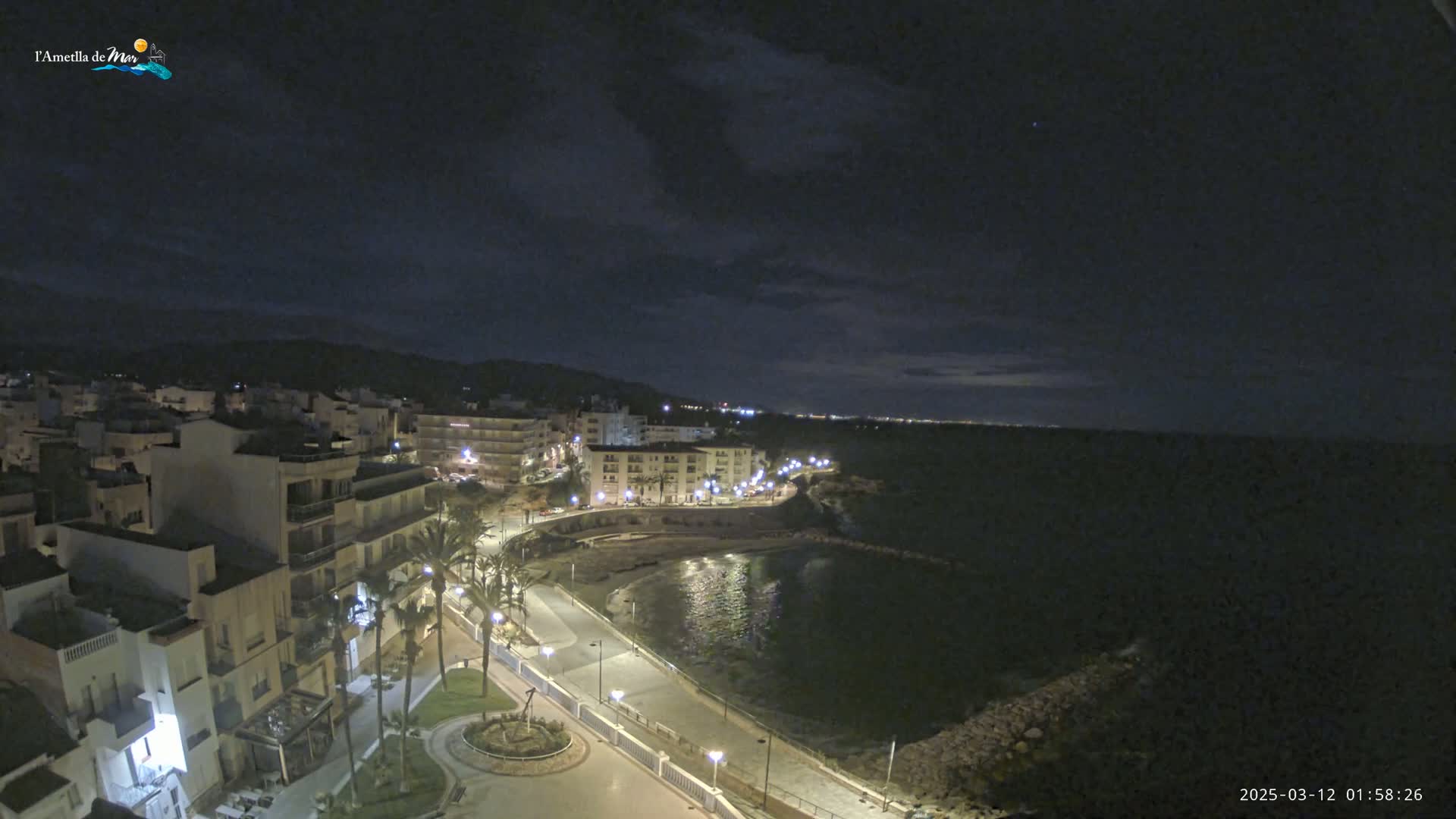 A nighttime, coastal town scene with a calm bay, illuminated buildings, and a partly cloudy sky.