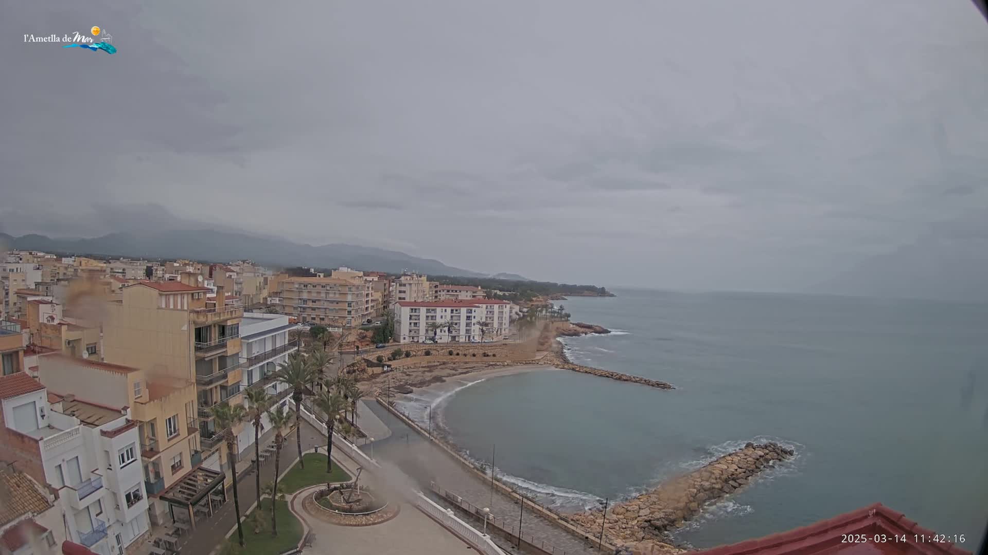 A coastal town with multi-story buildings, palm trees, and a small beach is seen under an overcast sky.