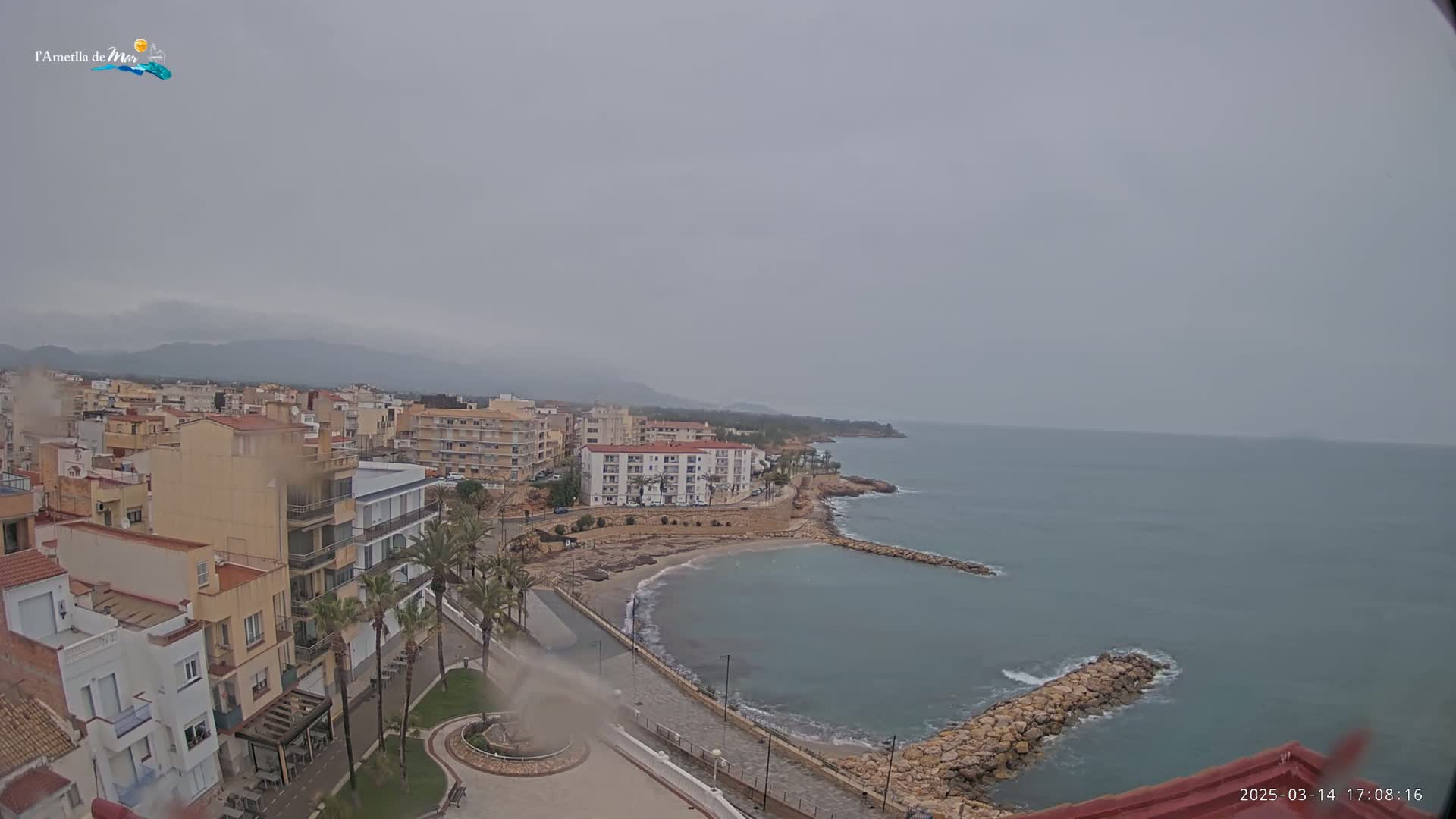 A coastal town with buildings, palm trees, and a small beach is seen on an overcast day.