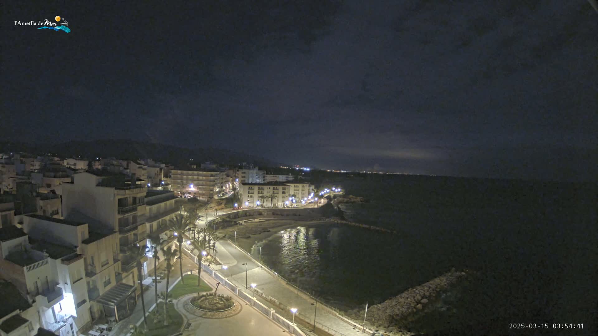 A nighttime, coastal town scene shows a crescent-shaped bay with a paved walkway lined with palm trees and buildings illuminated by streetlights under a dark, cloudy sky.