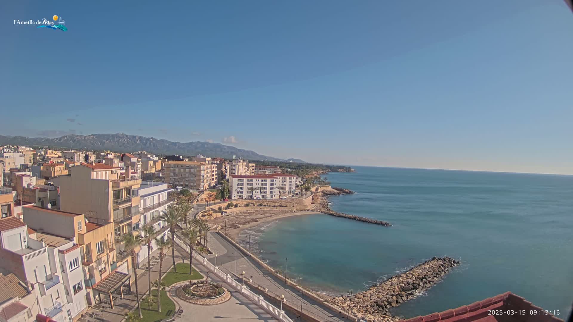 A coastal town with buildings and palm trees sits on a clear, sunny day next to a calm, blue ocean with rocky jetties.
