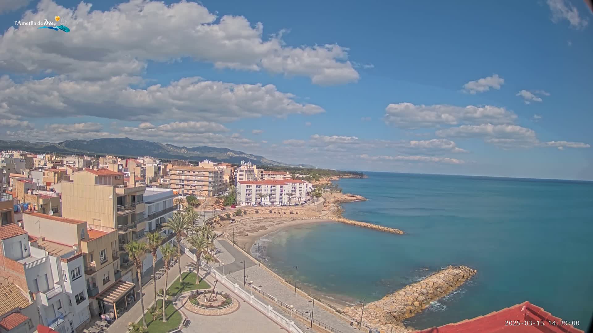 A coastal town with buildings and palm trees sits next to a calm, blue sea under a partly cloudy sky.