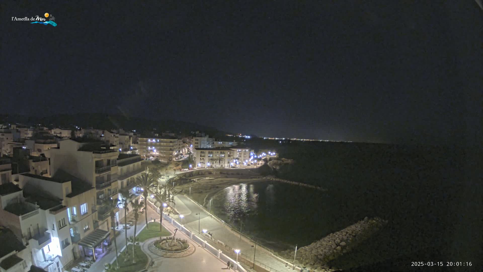 A nighttime, coastal town scene with calm waters reflecting the lights of buildings along a paved waterfront.