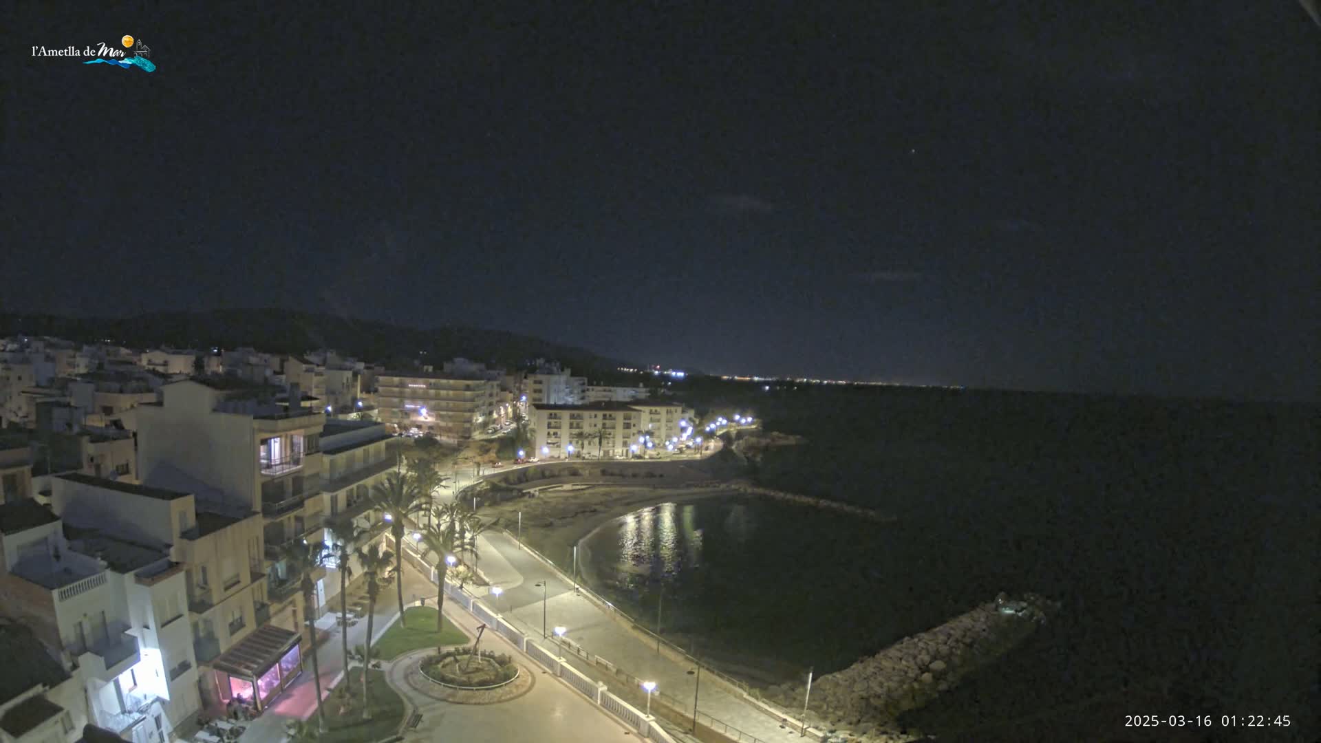A nighttime aerial view of a coastal town with a calm bay, illuminated buildings, and palm trees lining a paved walkway.