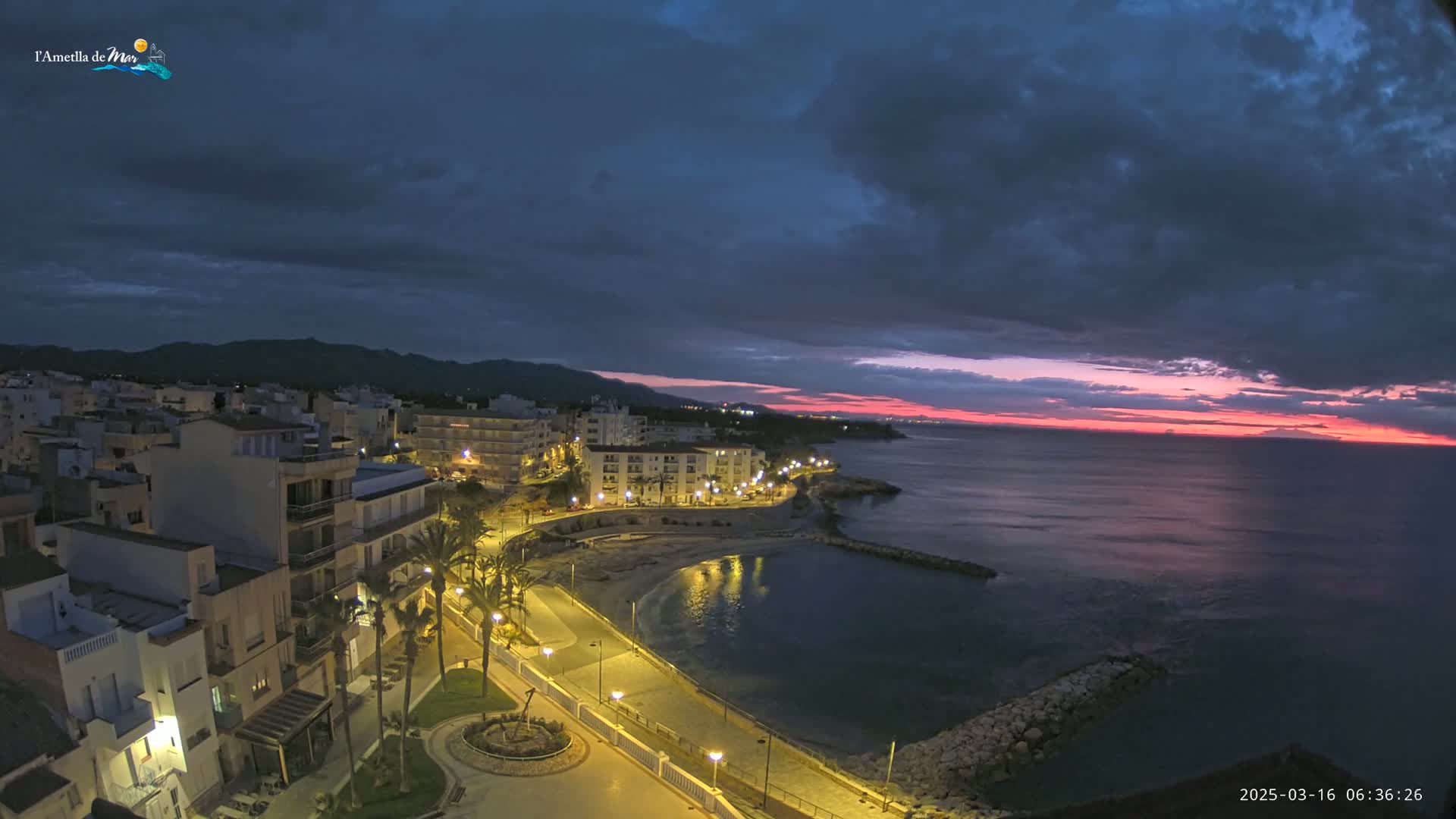 A coastal town at twilight, illuminated by streetlights, with a calm sea reflecting a pinkish sunrise under a partly cloudy sky.