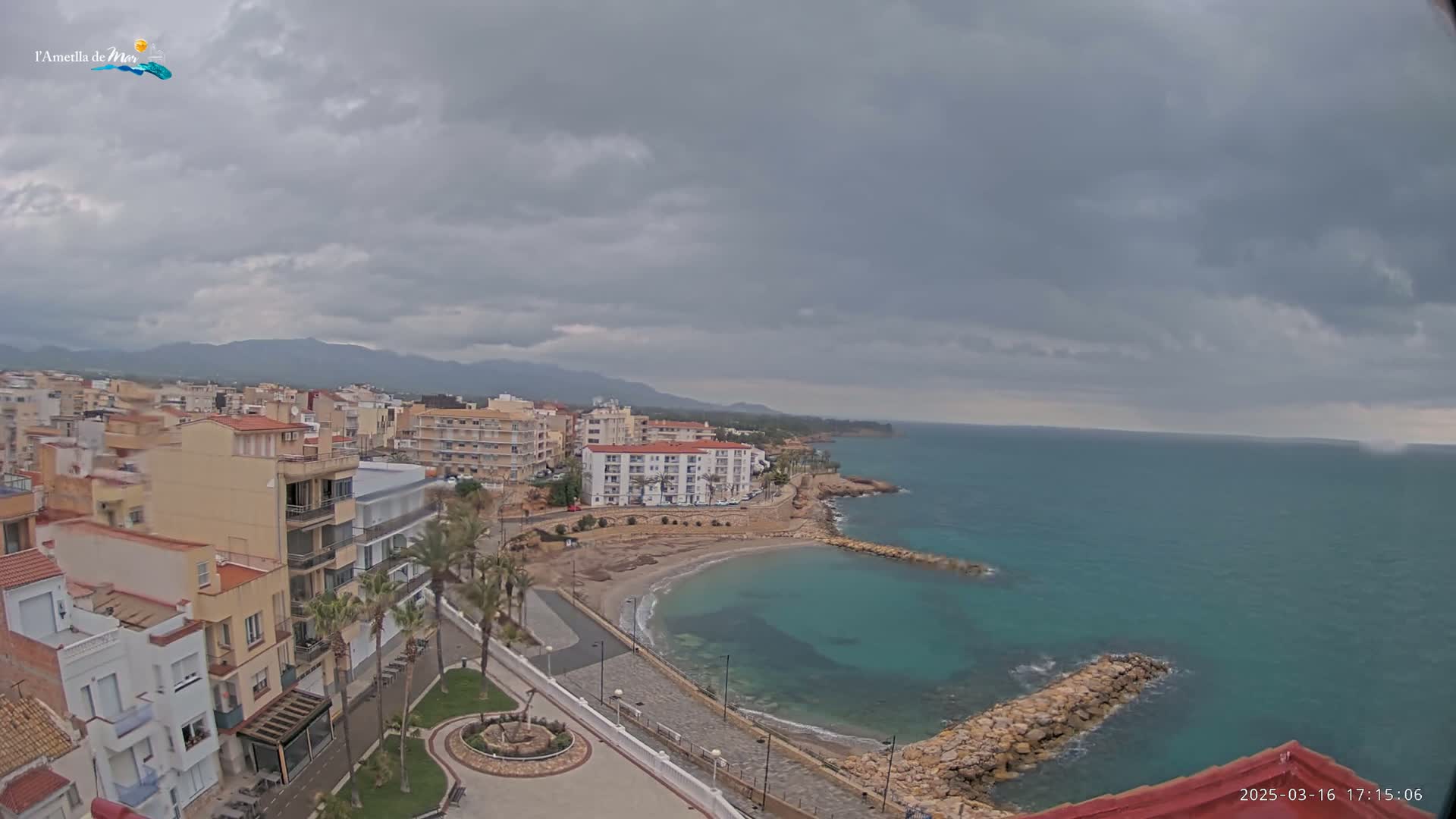 An overcast sky hangs over a coastal town with buildings clustered near a small, calm bay; a rocky breakwater extends into the teal sea.