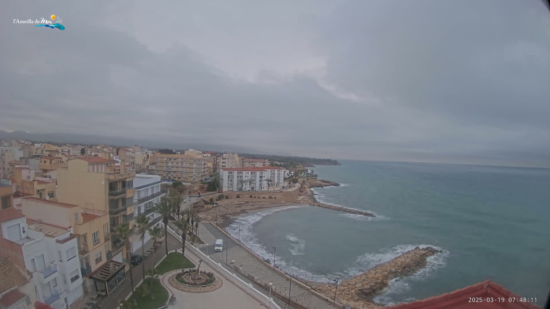 An overcast day reveals a coastal town with buildings lining a paved waterfront next to a calm sea with small waves.