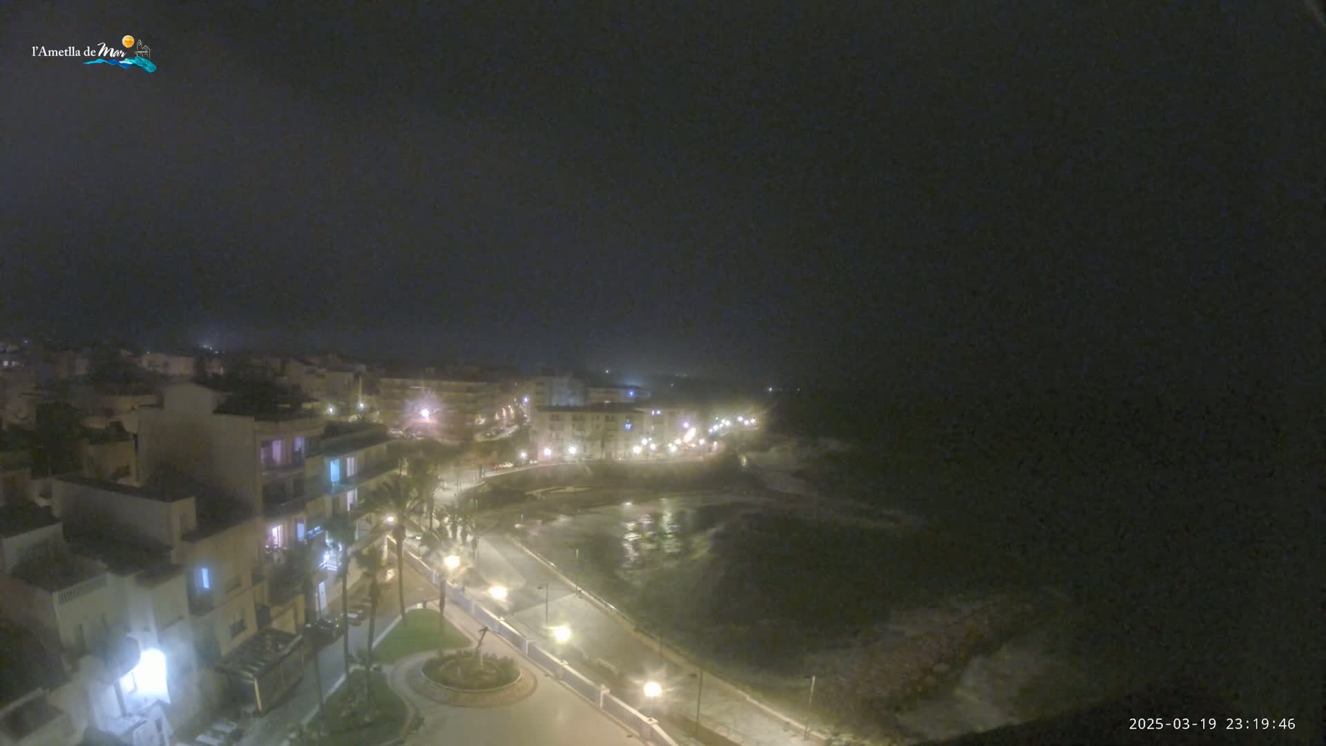 A nighttime, foggy view of a coastal town with illuminated buildings and streets leading to a dark, wavy ocean.
